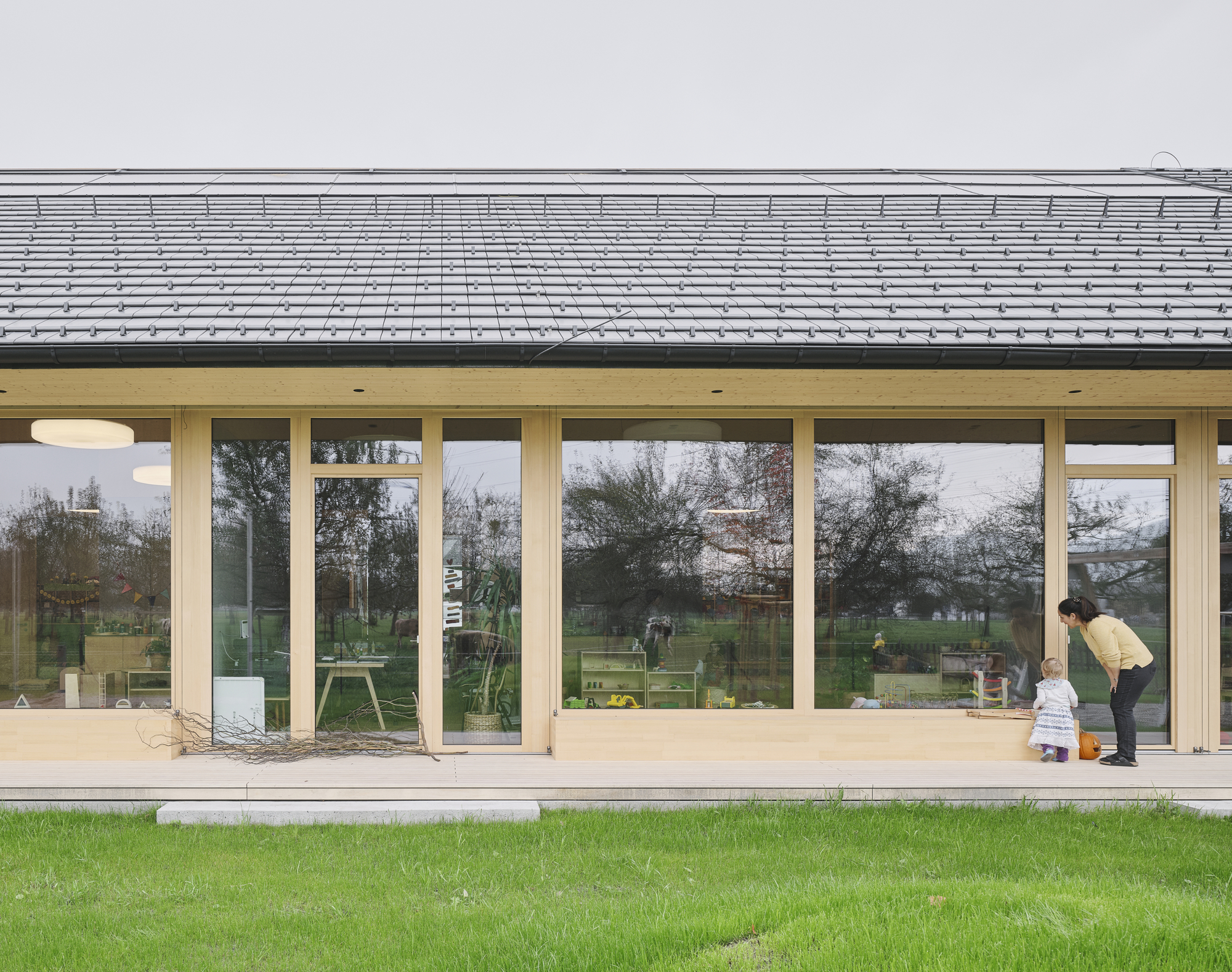 Glazed facade with timber mullions beneath roof overhang showing figure bending near entrance