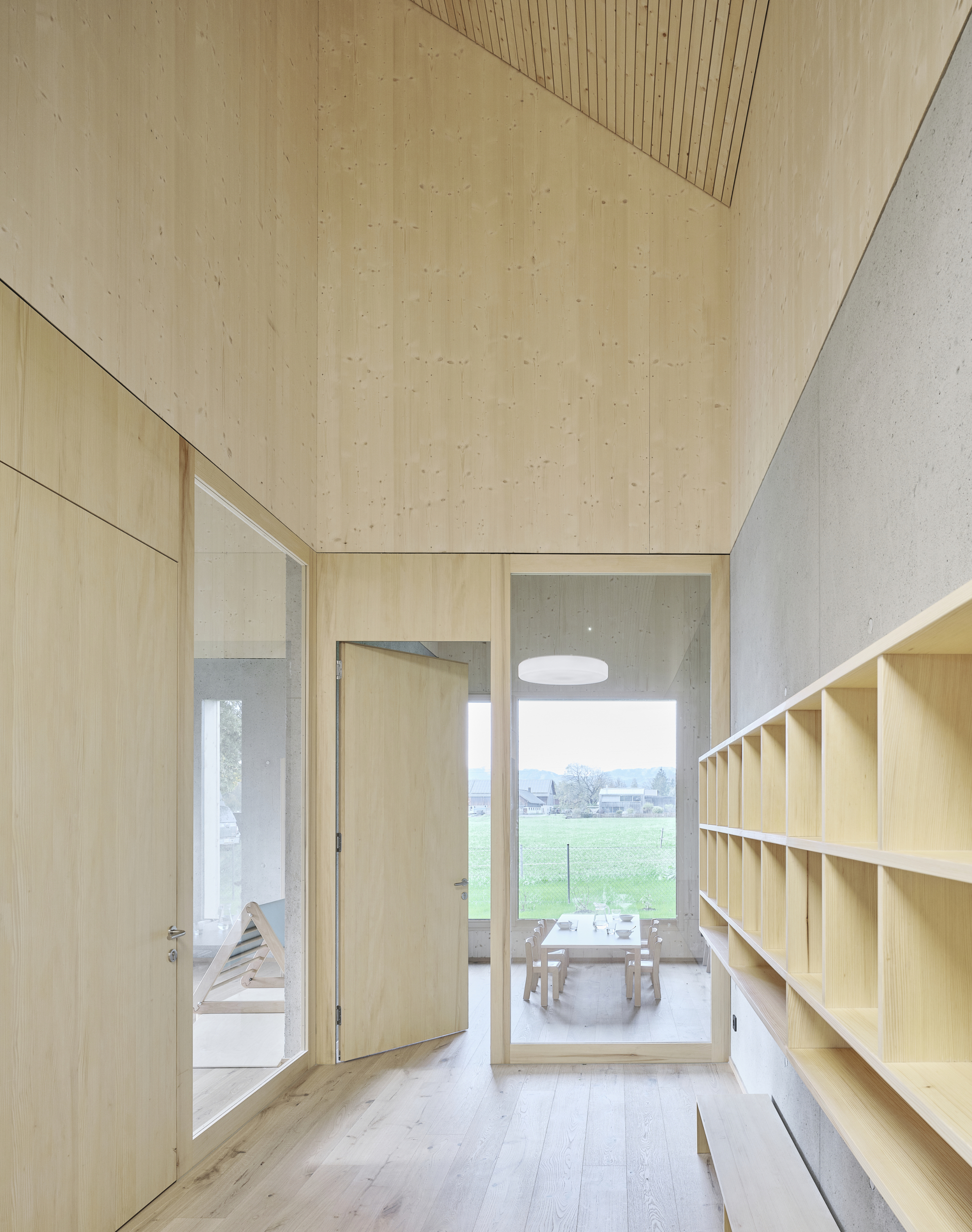 Corridor with plywood walls and slatted ceiling looking through open doors to a dining area beyond the orchard
