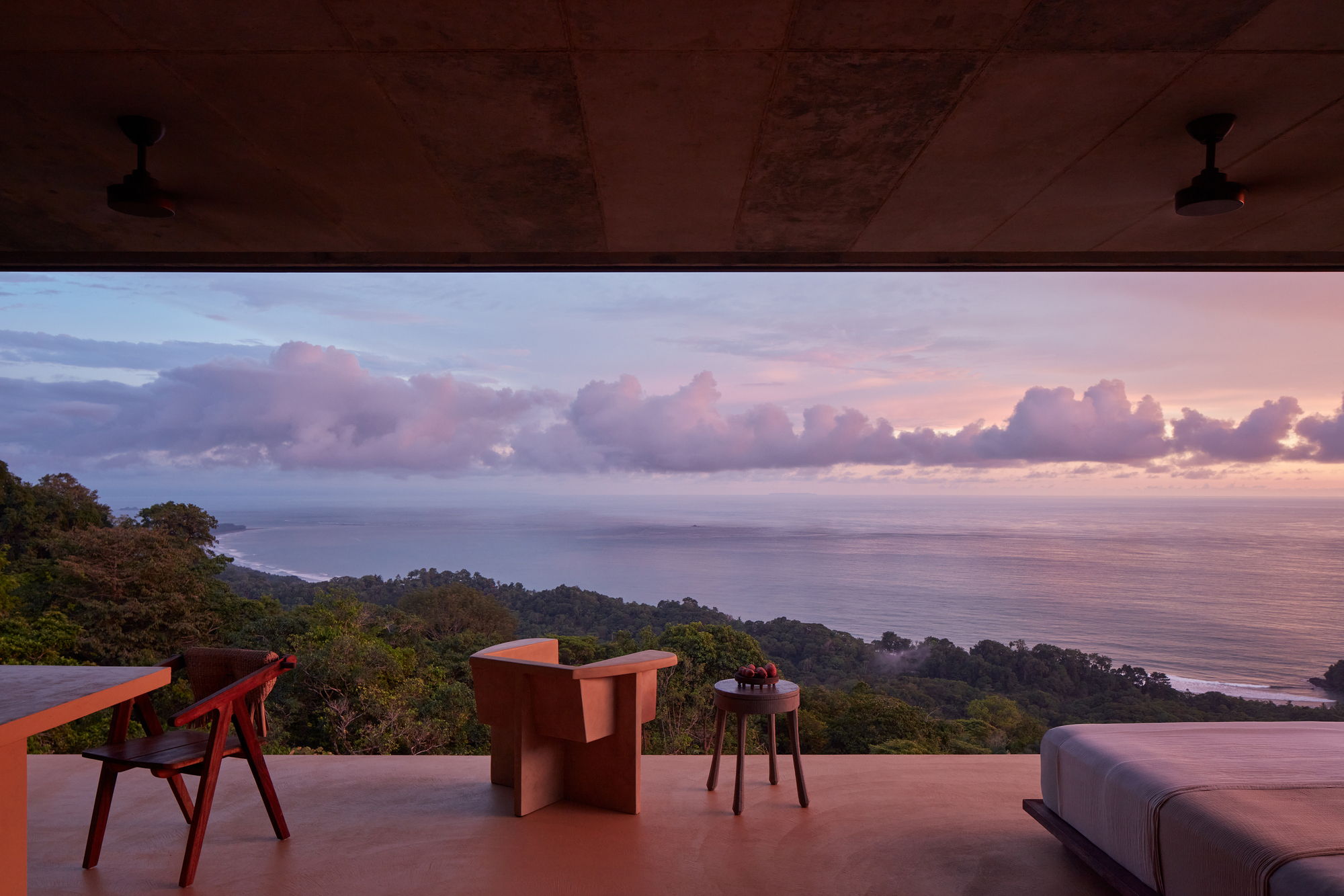 Open-air living space with red concrete floor overlooking forested hillside and ocean at sunset