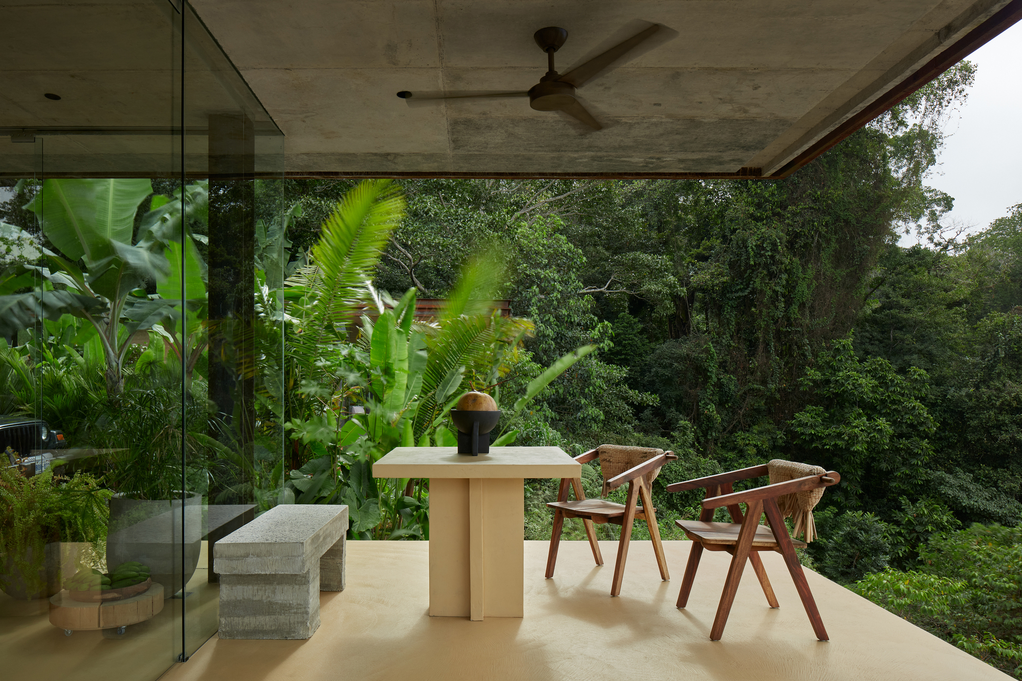Covered dining terrace with exposed concrete ceiling and banana plants framing forested view