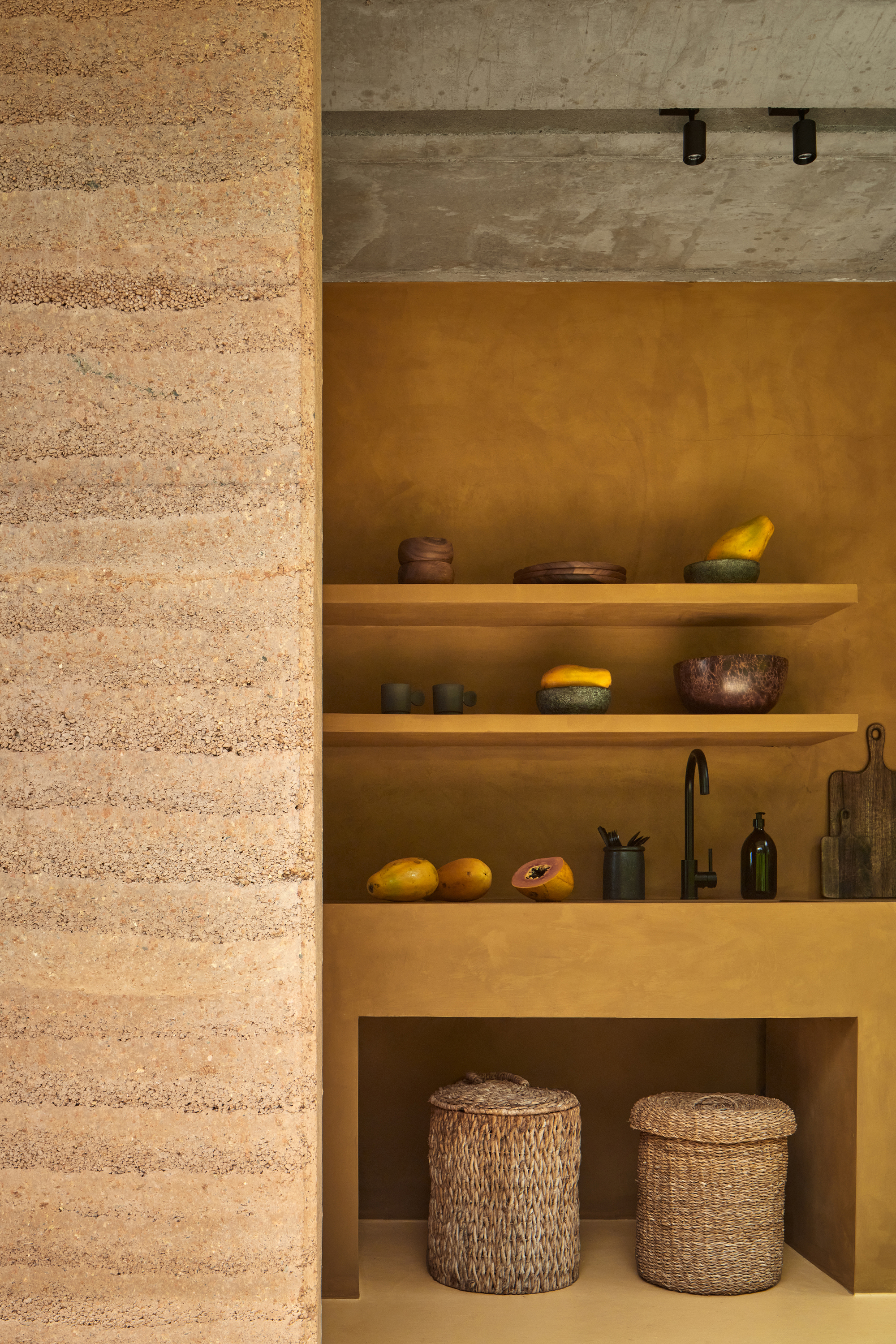 Kitchen alcove with floating timber shelves against ochre plaster walls beside a rammed earth partition