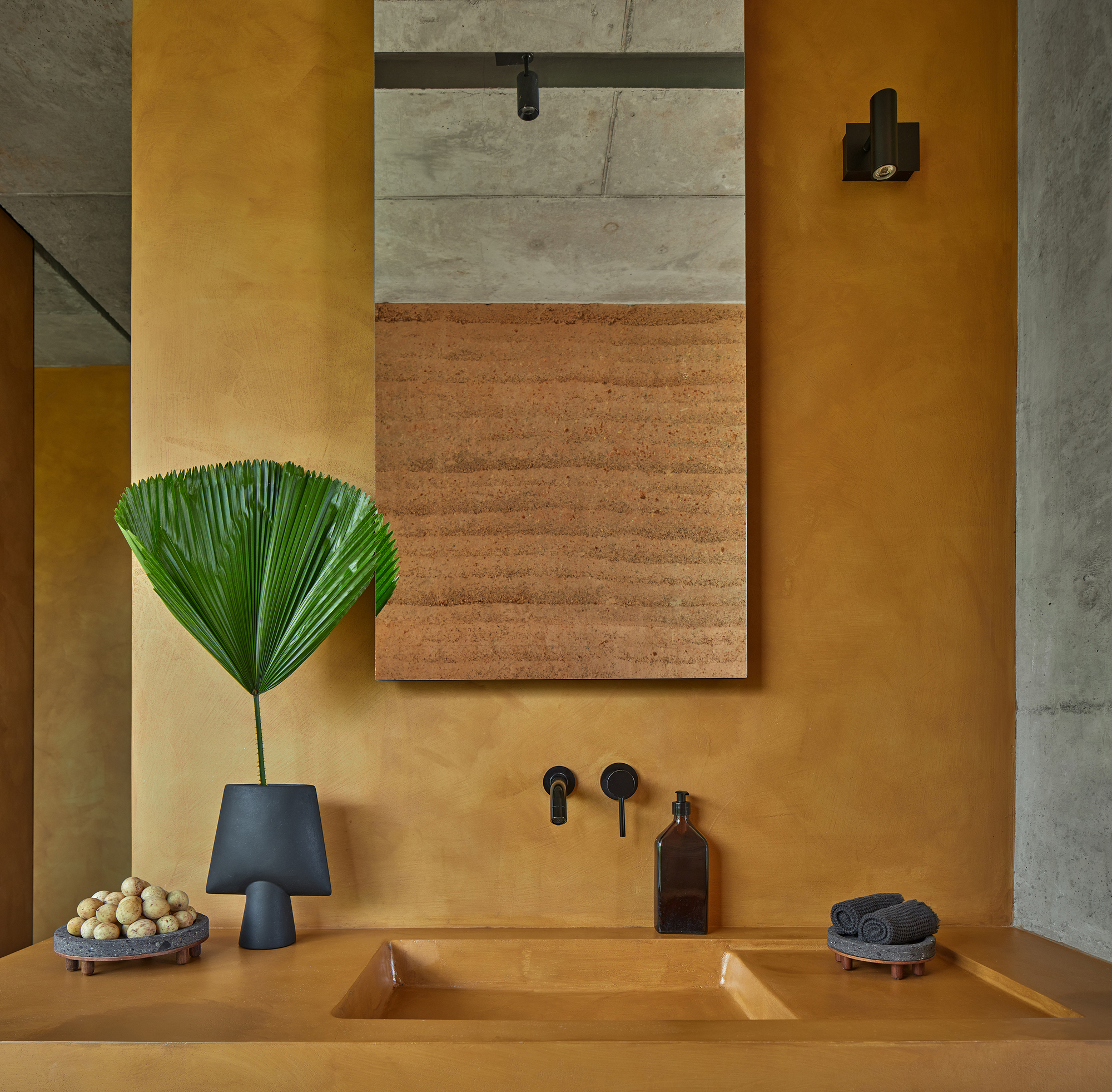 Bathroom sink detail with black tapware and framed rammed earth panel above ochre plaster countertop