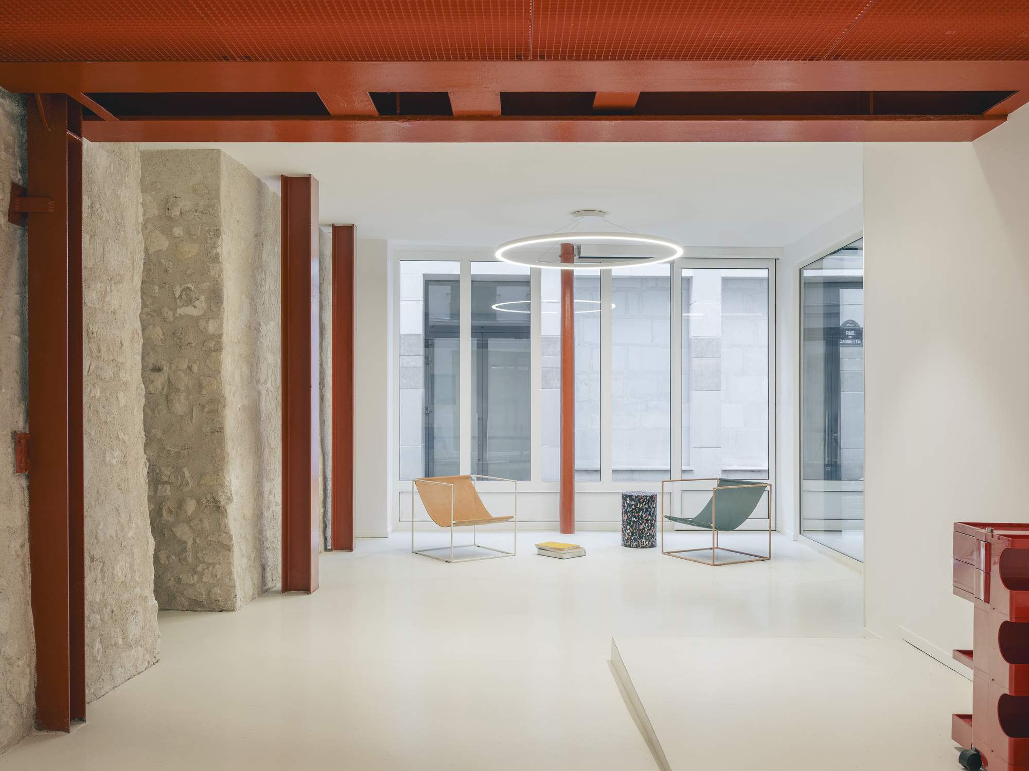 Interior space with exposed stone walls, red steel columns, perforated metal ceiling and glazed courtyard beyond