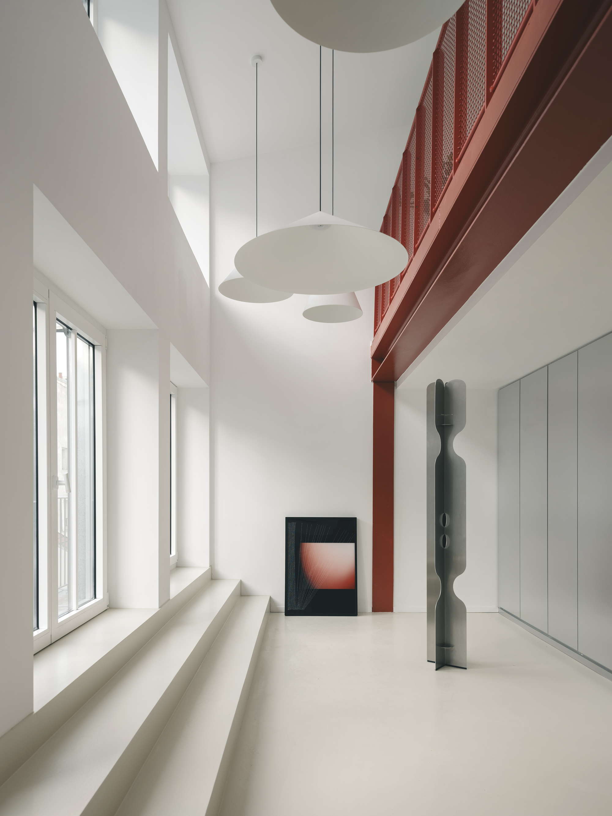 Double-height interior with red perforated metal mezzanine, pendant lights, and sunlight through tall windows