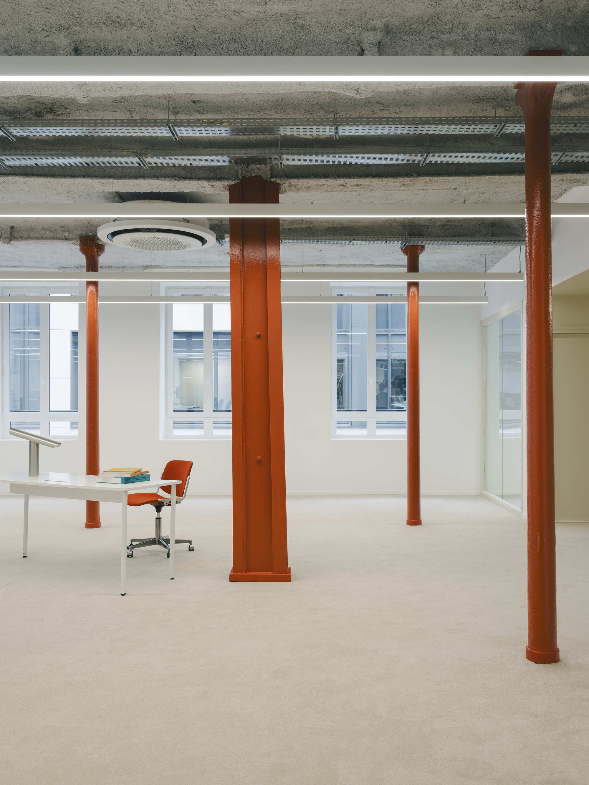 Orange steel columns framing workspace with desk and chair beneath exposed concrete ceiling and ducting