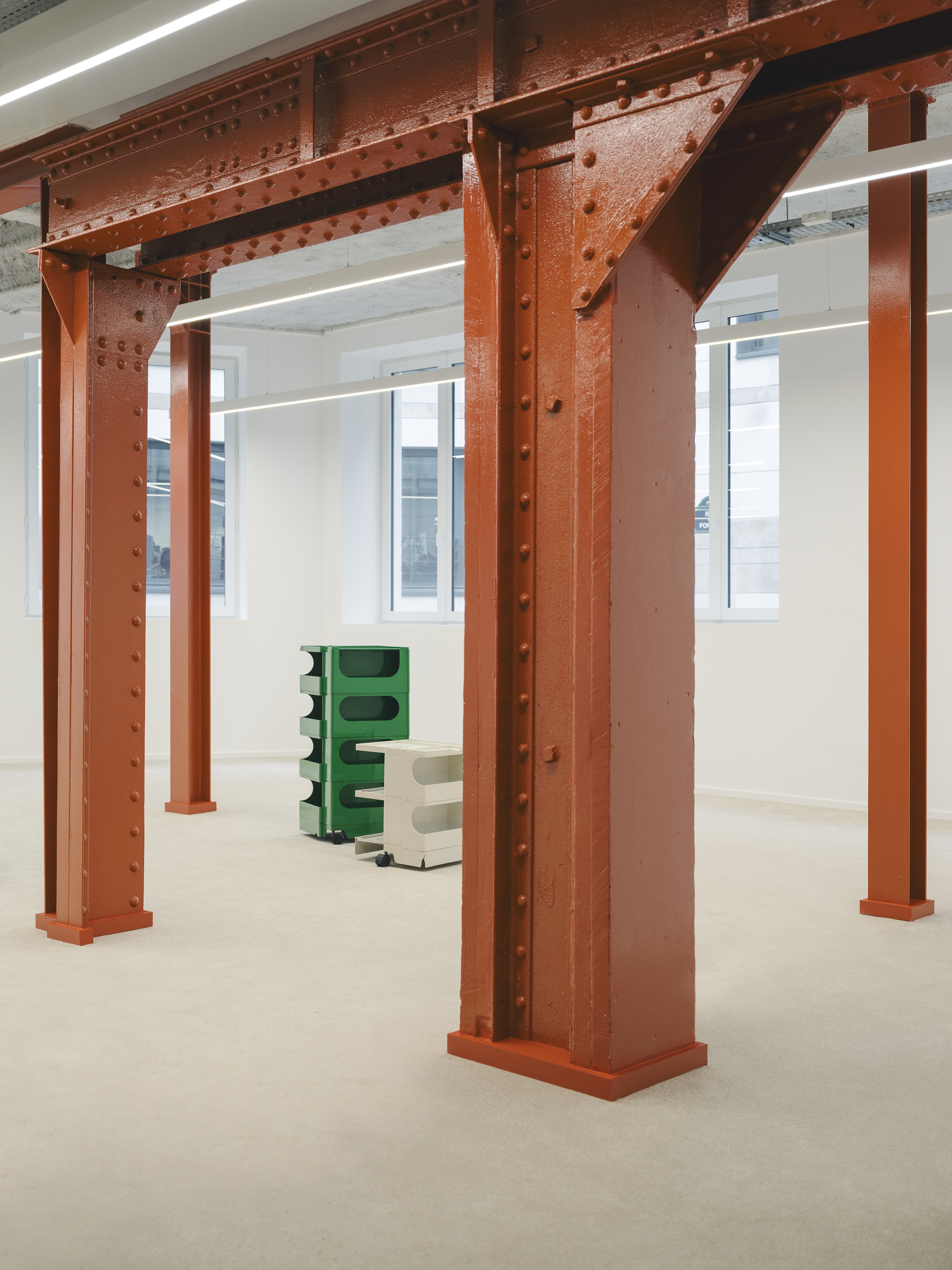 Exposed riveted steel beams and columns painted red oxide in an open floor space with white walls