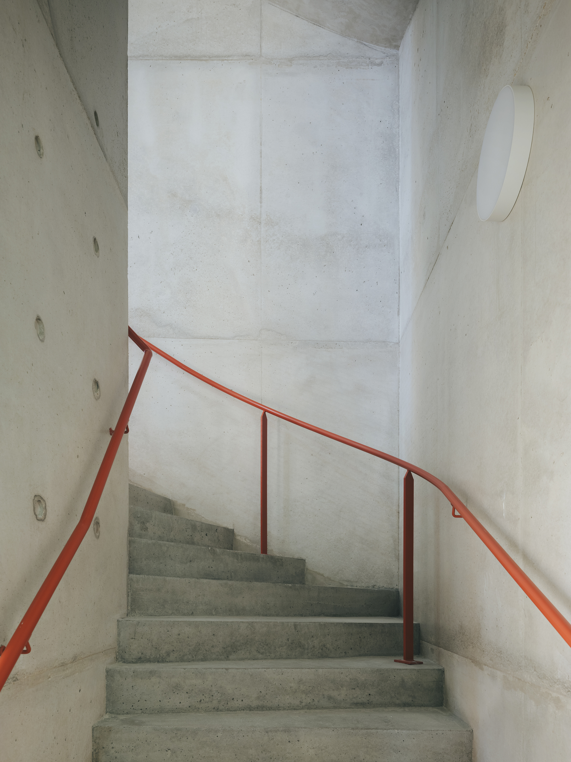 Concrete stairwell ascending between curved walls with a red handrail following the turn