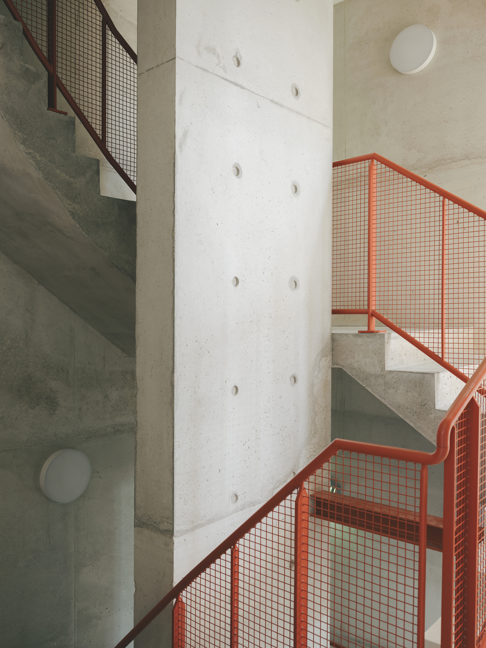 Concrete staircase with orange mesh railings winding around a cylindrical column with circular apertures