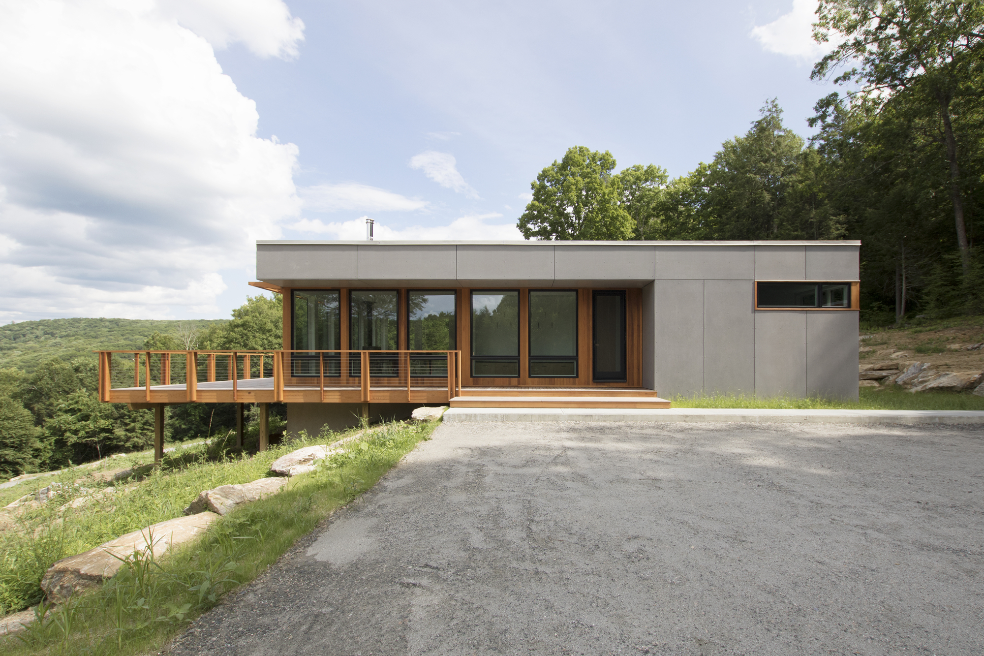 Street-facing view of the gray panel facade with cantilevered timber deck overlooking a forested hillside
