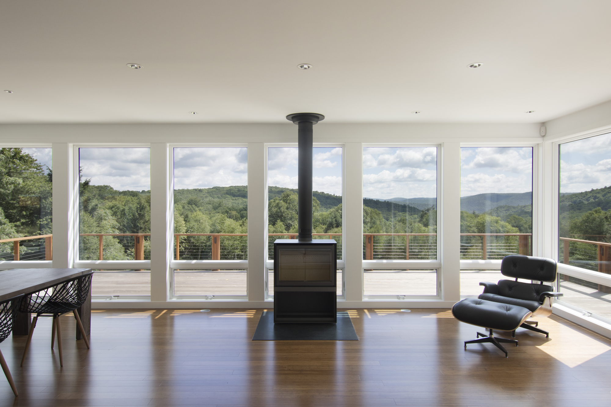 Open living space with freestanding black stove and full-height glazing facing forested hills
