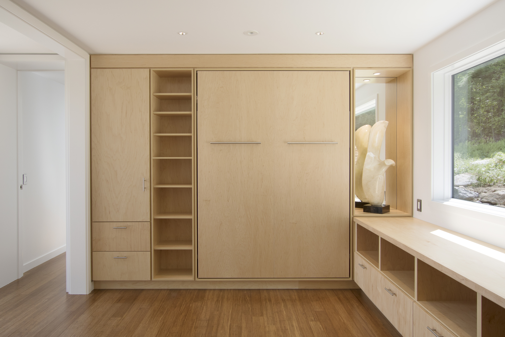 Light wood wall of built-in storage with murphy bed and shelving beside a window with garden view