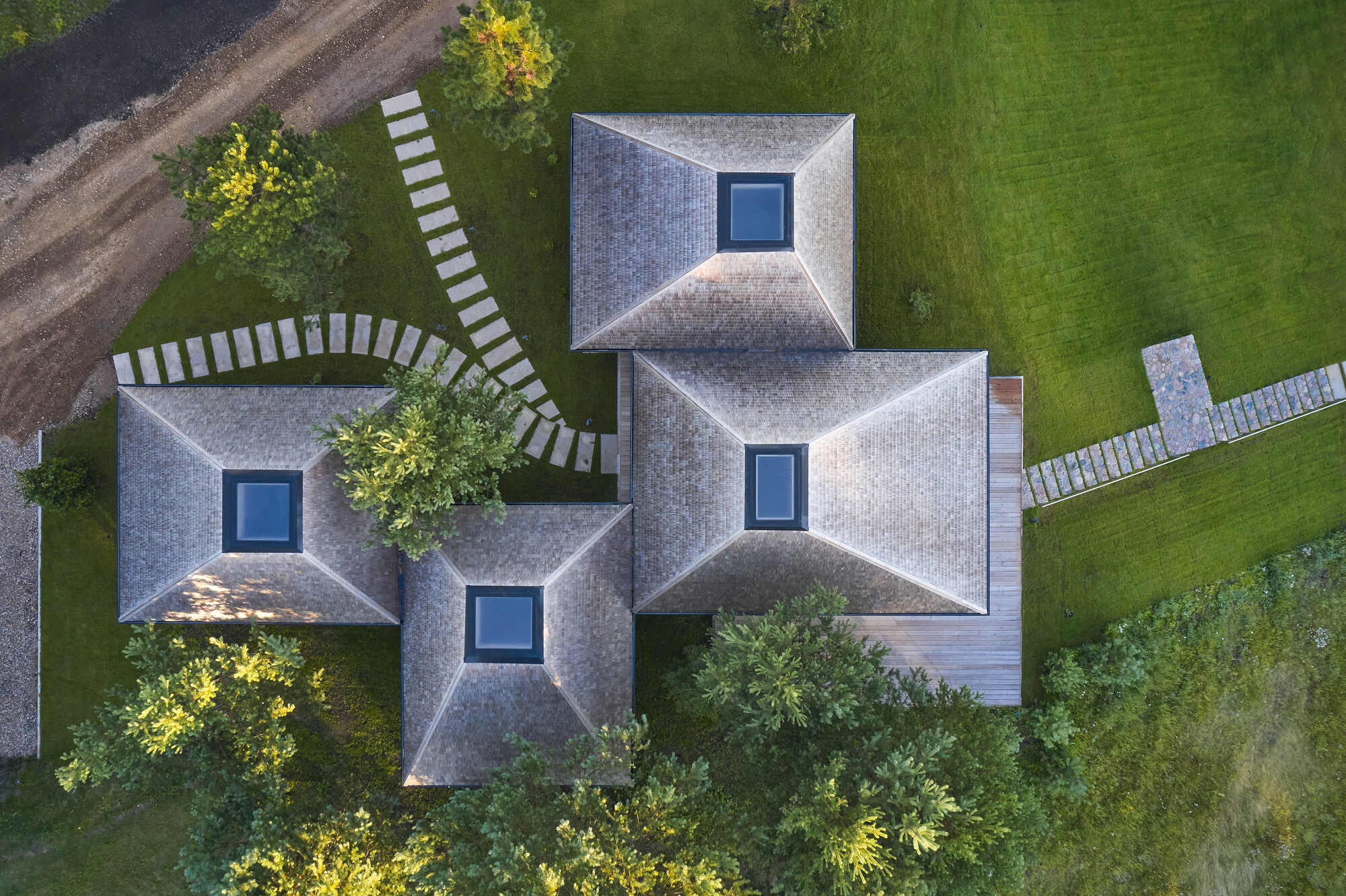 Drone view of four pyramidal shingled roofs with central skylights arranged around planted courtyards and stone pathways