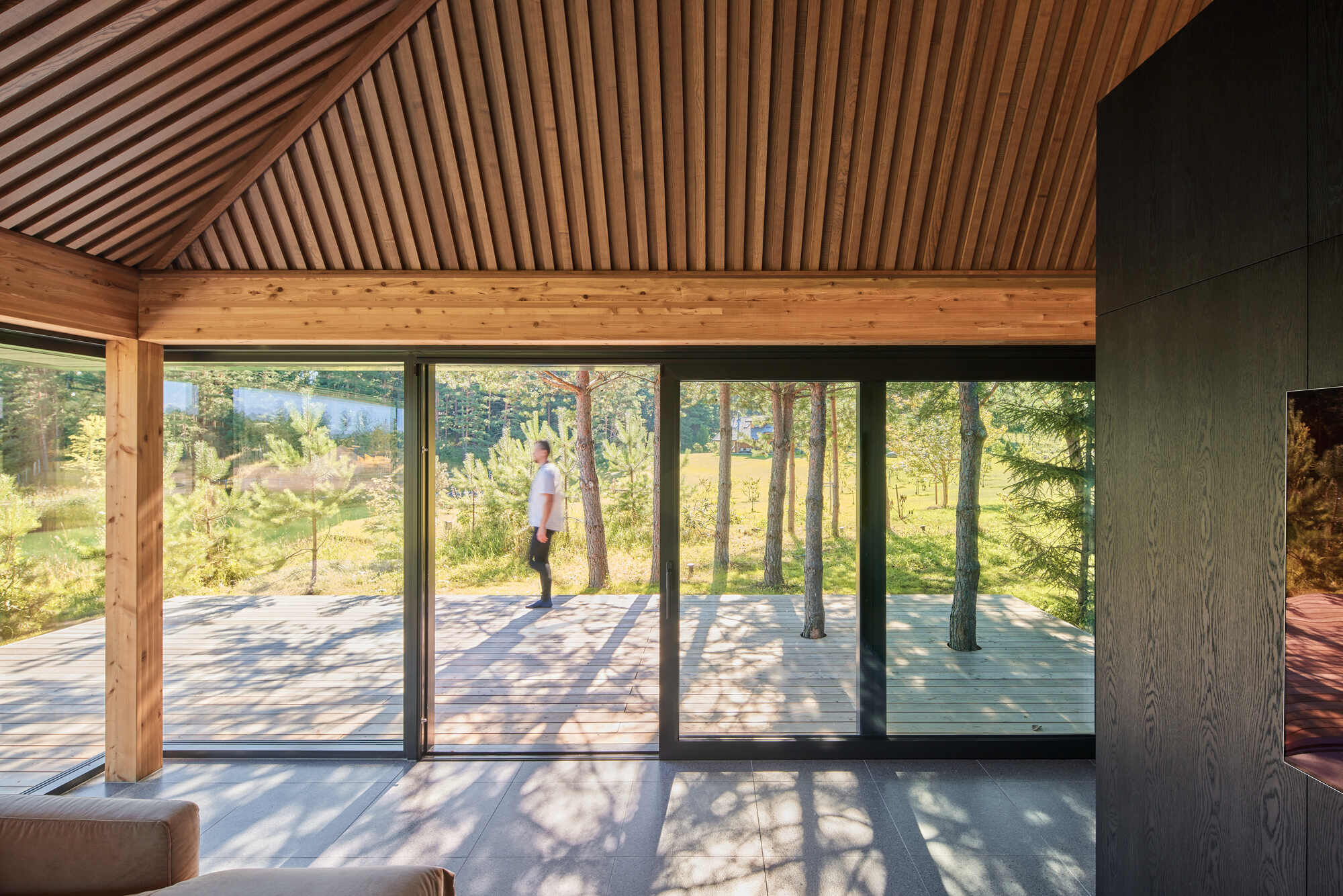 Interior view through floor-to-ceiling glazing to exterior terrace with timber slat ceiling above and figure standing outside