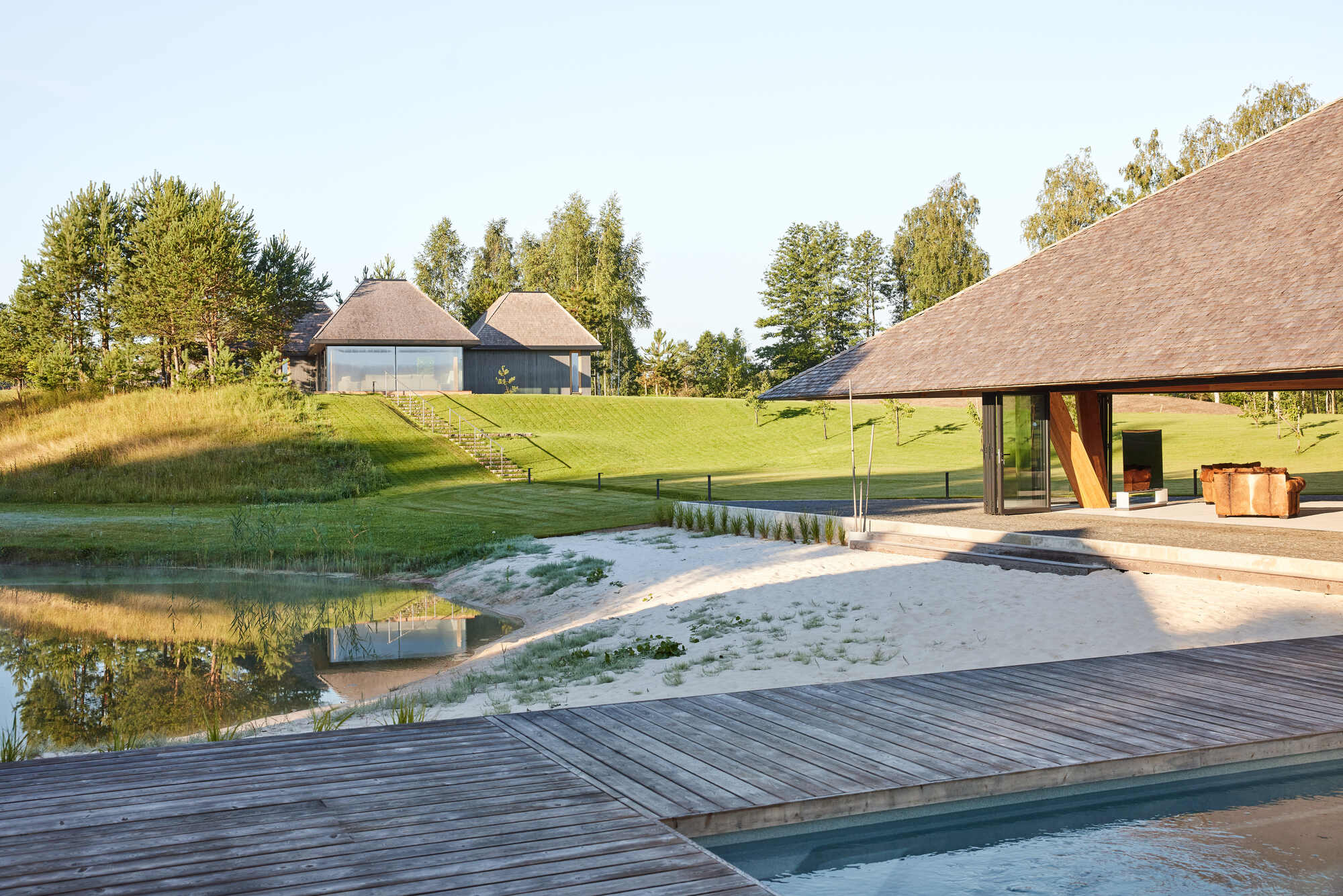 Timber deck and reflecting pool leading to glazed pavilion under a sloped roof in afternoon light