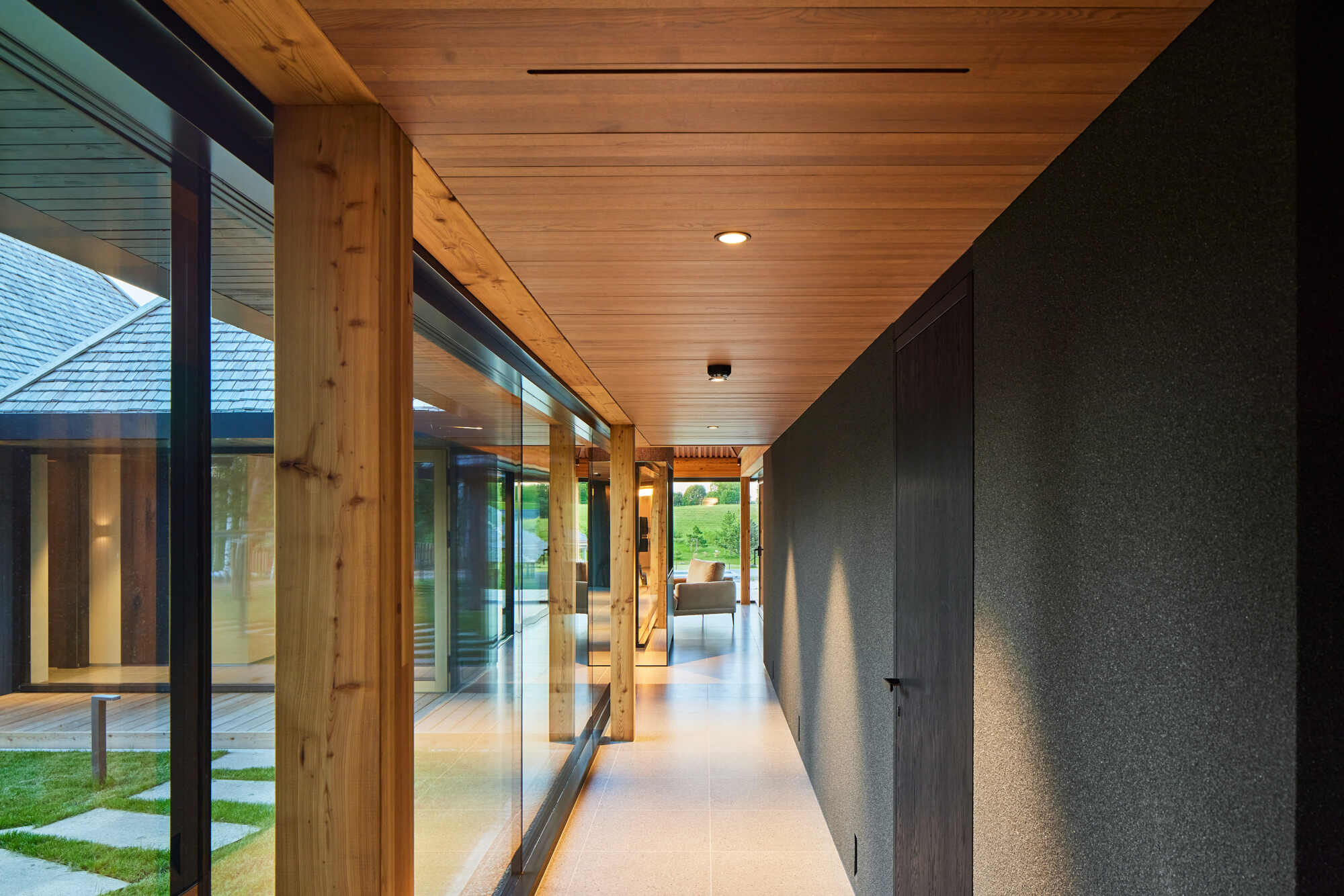 Glazed hallway with timber posts and plank ceiling looking toward another pavilion at dusk