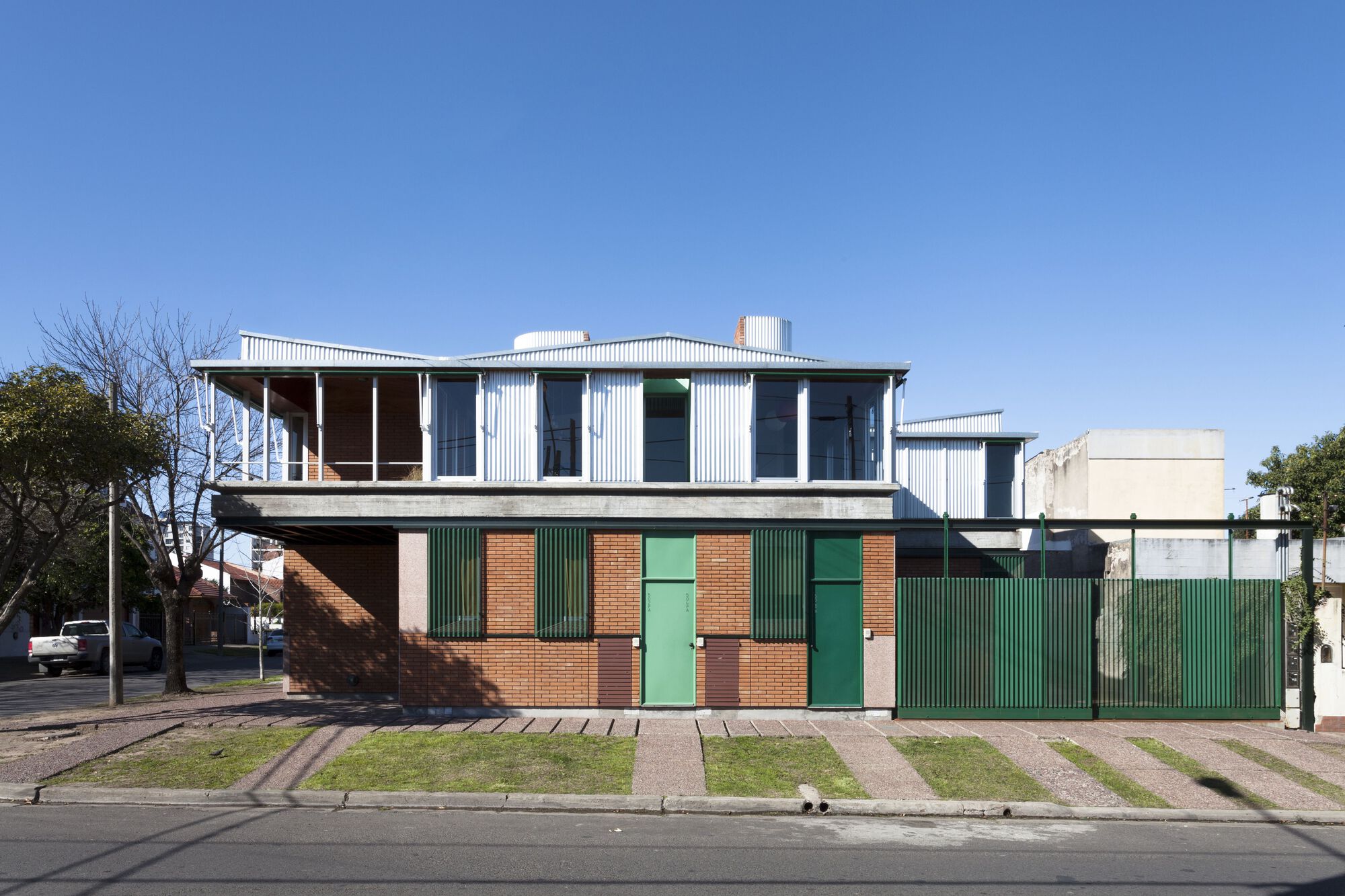 Street view of the two-story residence with red brick base, green corrugated shutters and white corrugated upper level