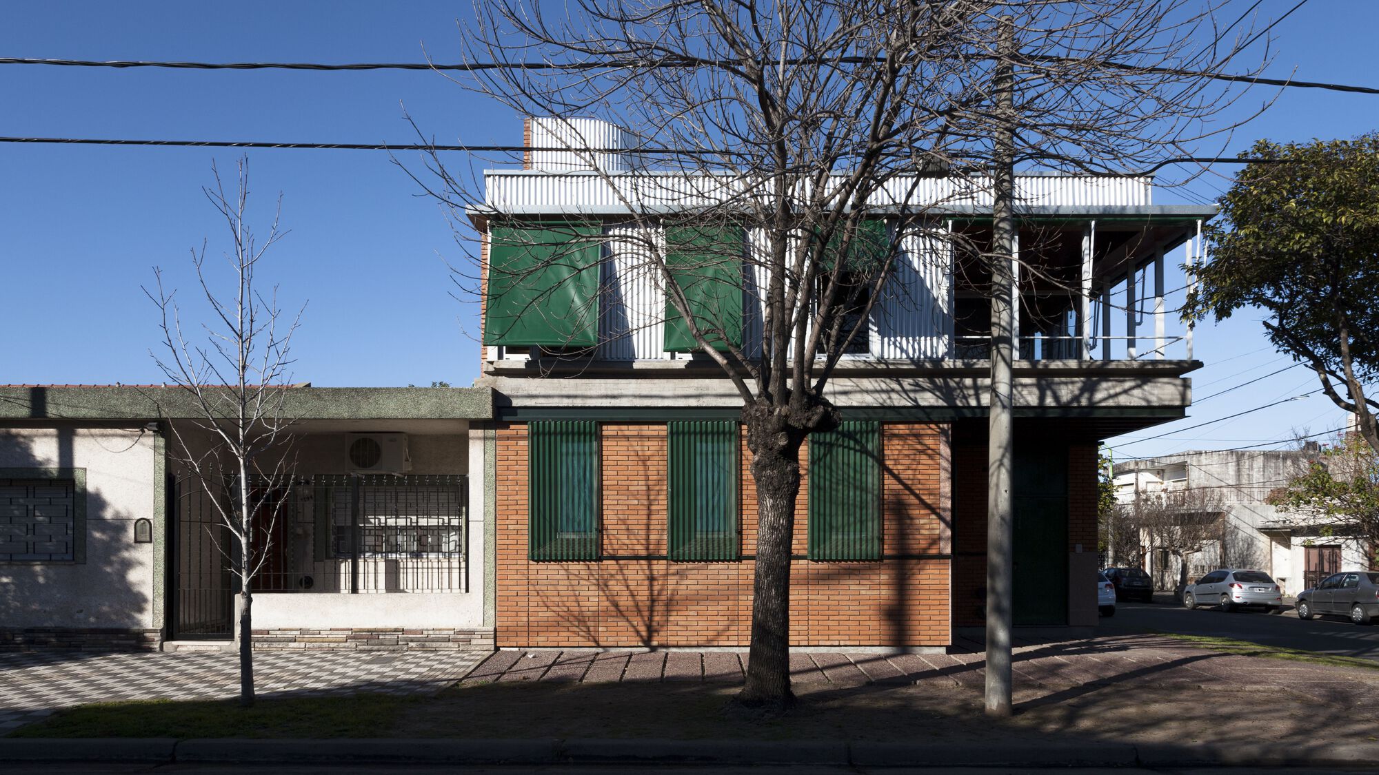 Corner elevation showing brick walls with green metal shutters and elevated white metal upper volume behind bare trees