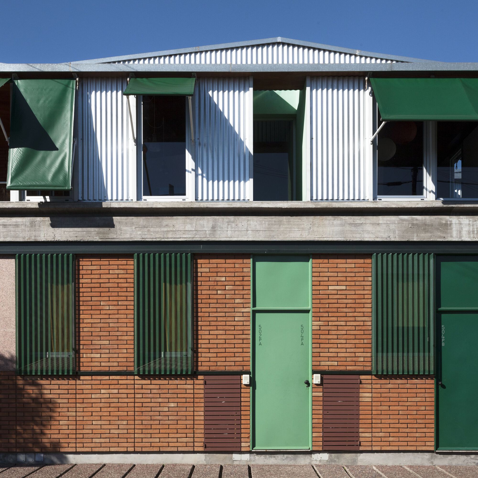 Two-storey facade with green corrugated metal shutters, orange brick panels, and white corrugated metal upper level under clear sky