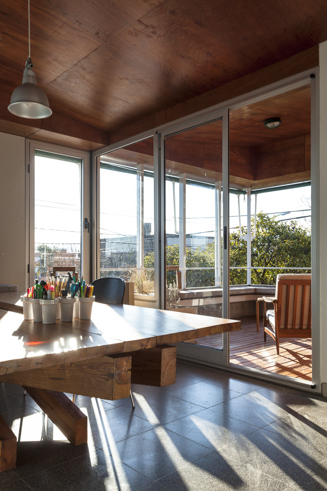 Timber dining table beneath plywood ceiling with sliding glass doors opening to timber deck and urban rooftops beyond