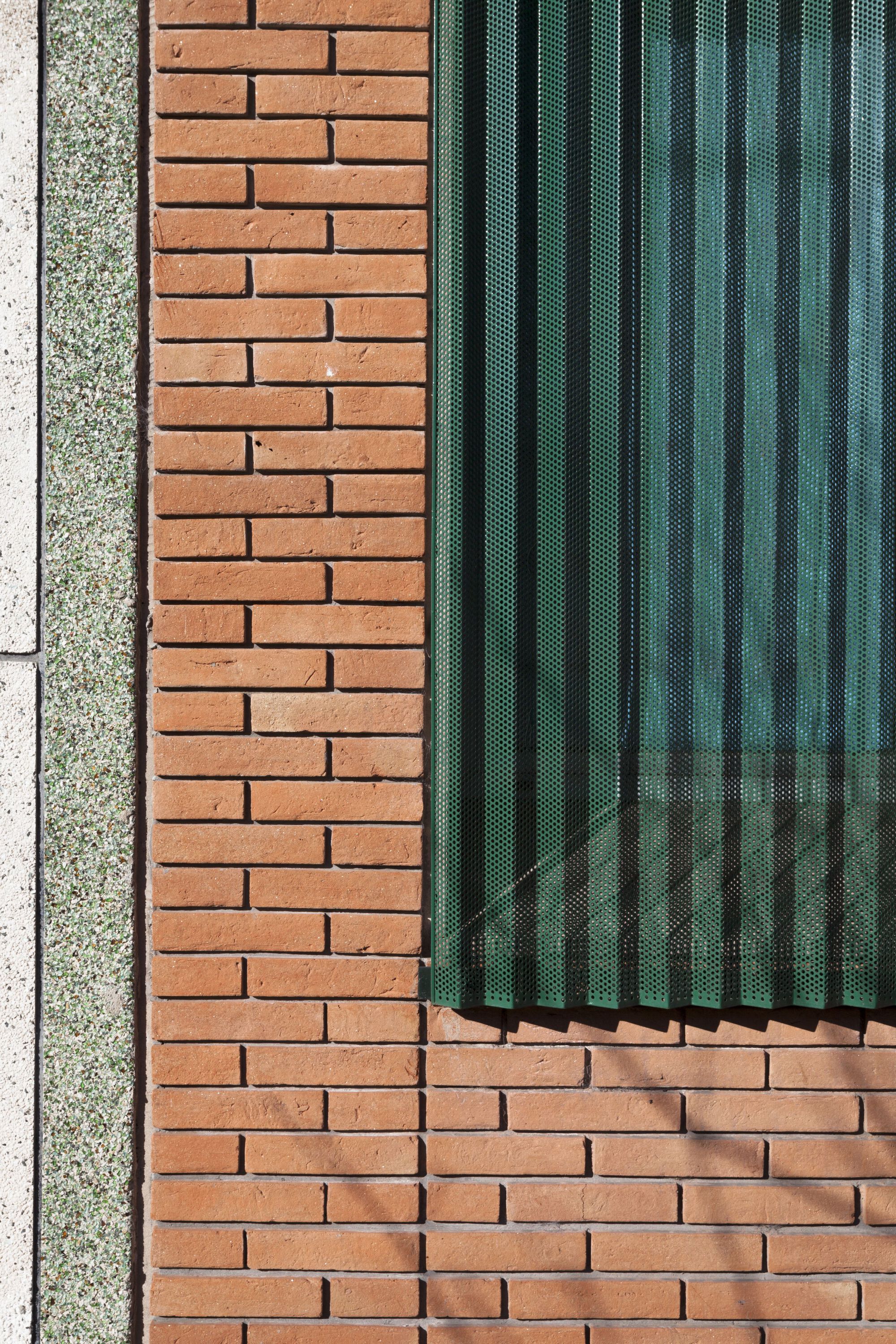 Detail of perforated green metal shutters meeting red brick wall with exposed aggregate concrete edge
