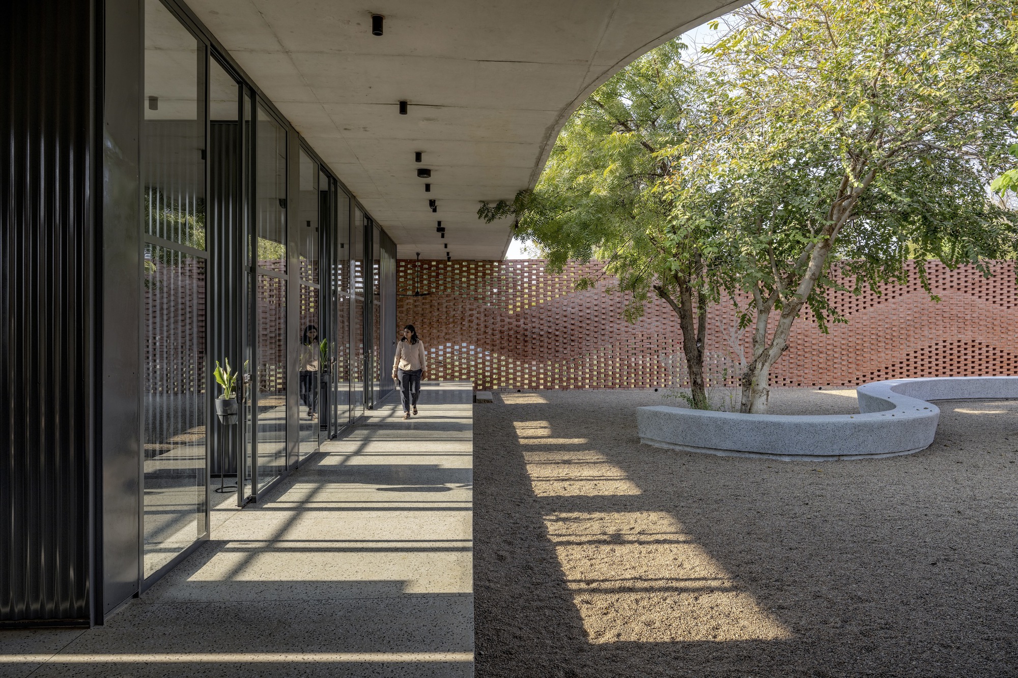 Covered walkway with vertical metal fins casting striped shadows onto the concrete floor and courtyard beyond