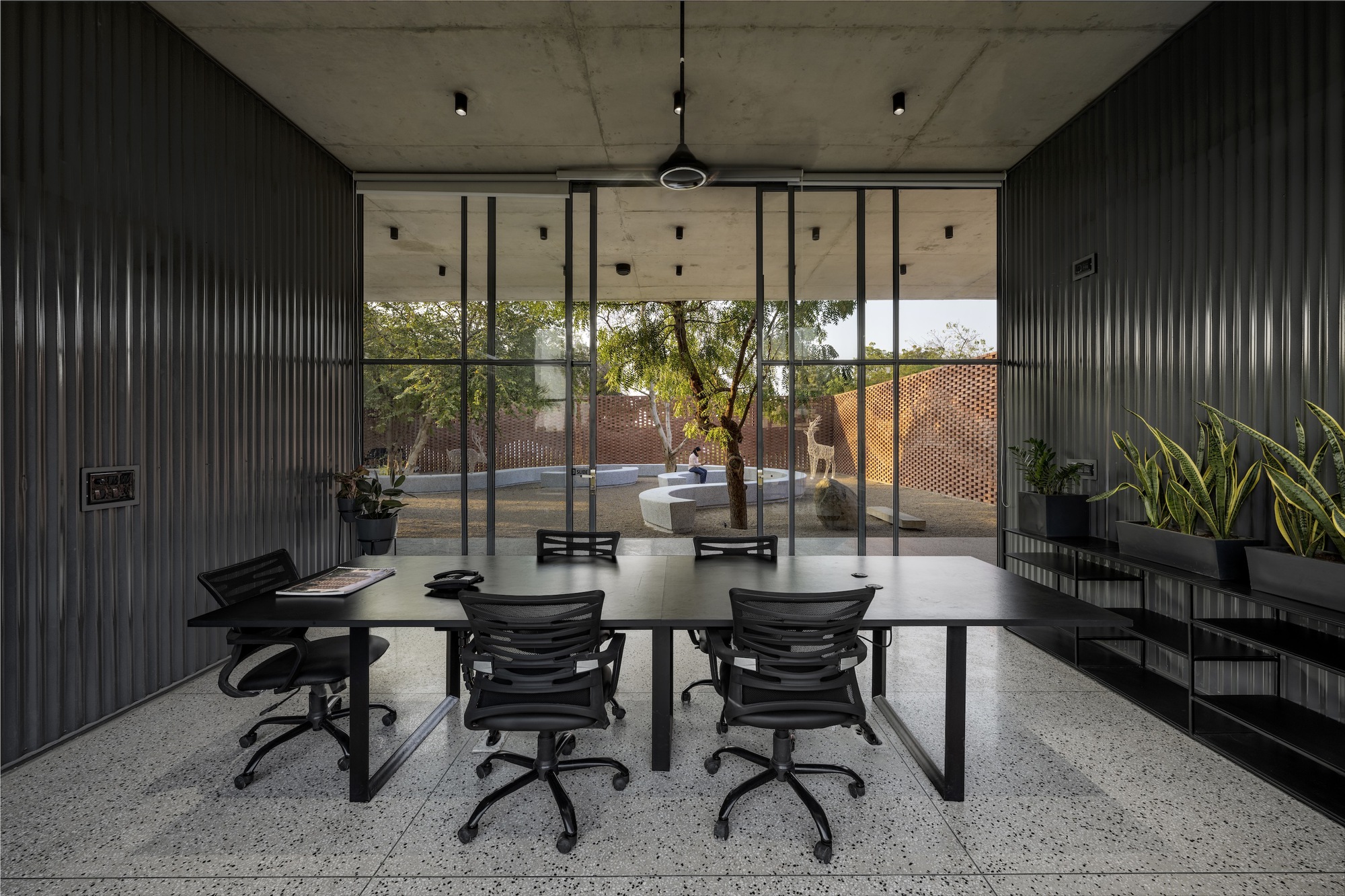 Conference room with black corrugated metal walls and floor-to-ceiling glazing opening to a courtyard with trees