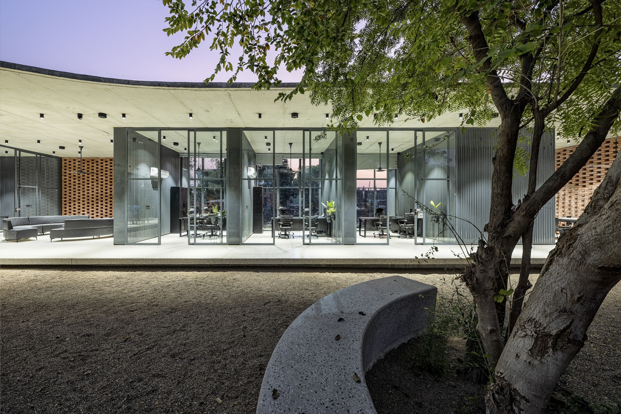 Glass facade beneath concrete overhang with existing mature tree shading a carved stone bench in gravel courtyard