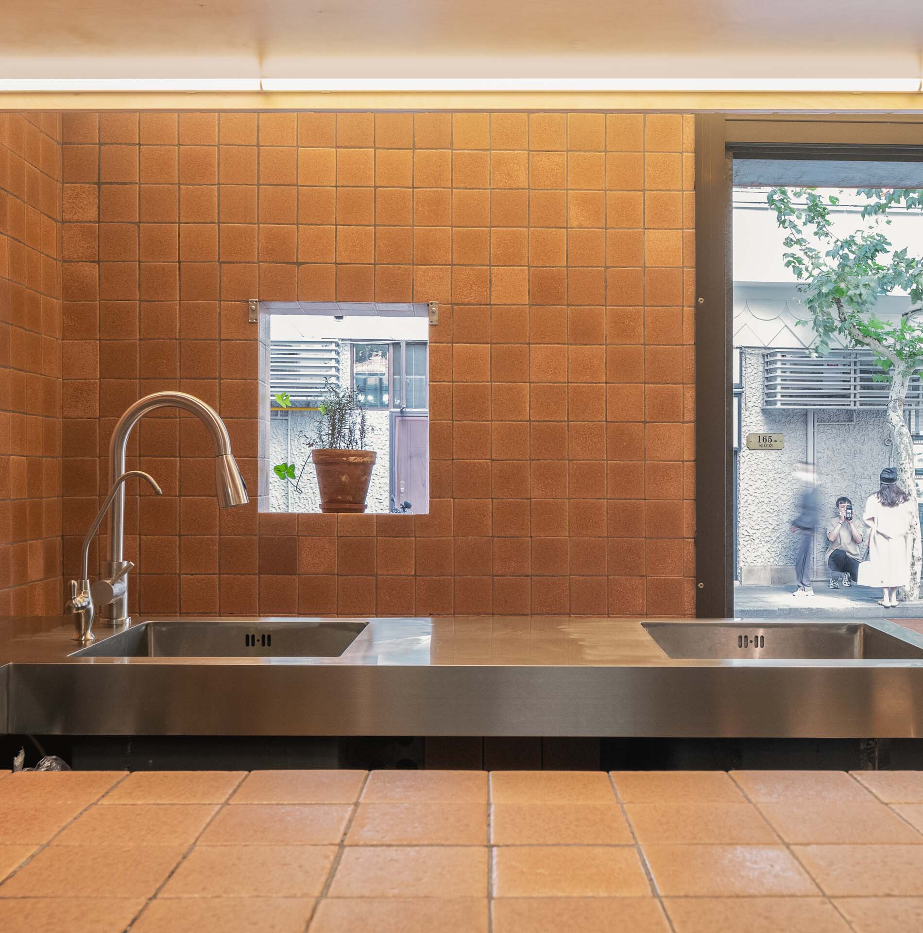 Stainless steel counter with dual sinks below orange tiled wall and storefront window showing pedestrians