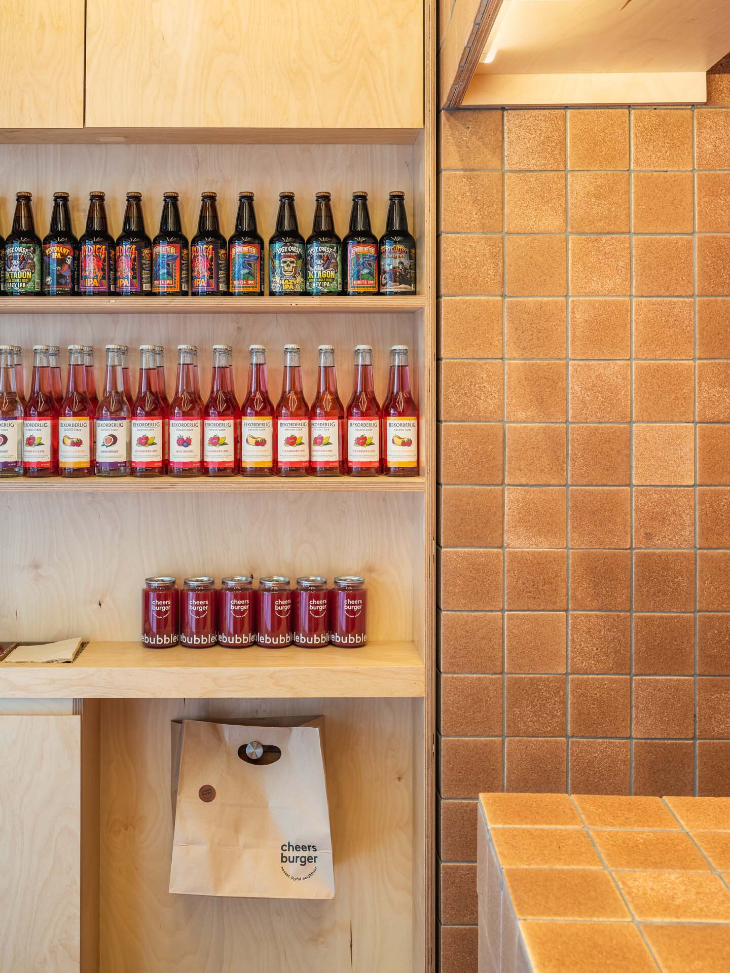 Open plywood shelving with bottled drinks beside orange tiled wall and takeaway bag below