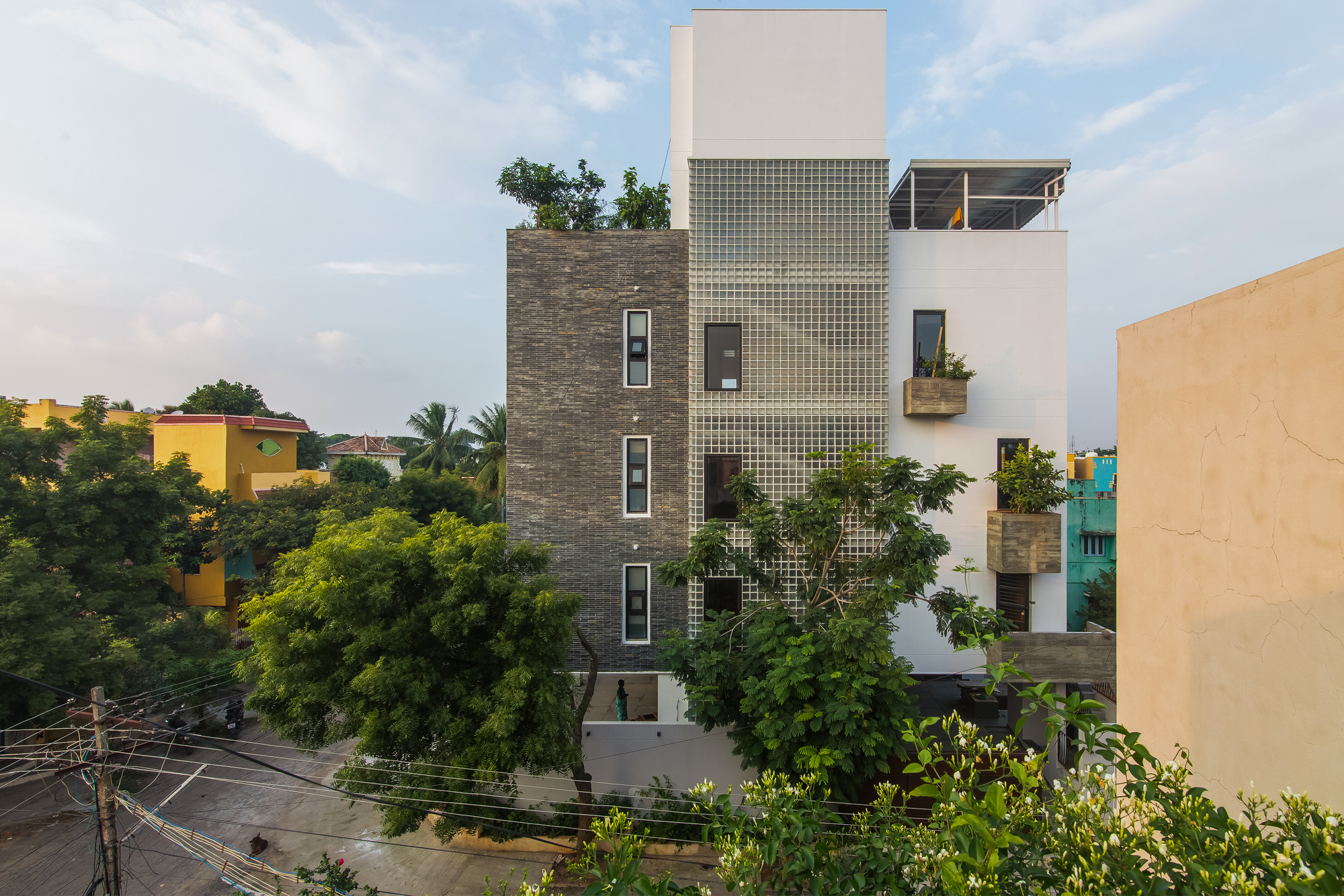Street facade combining stacked stone, glass block panels, and white stucco volumes with planted balconies