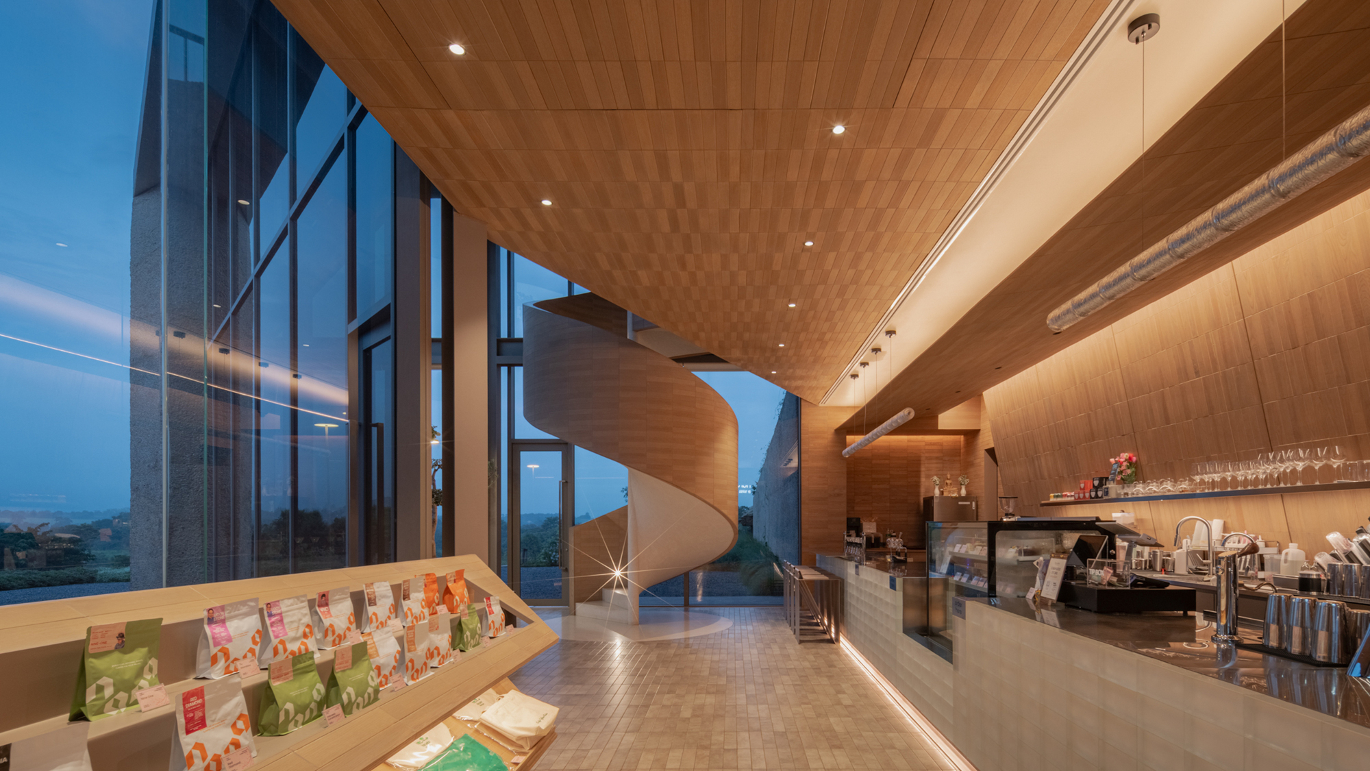 Double-height cafe space with curved timber ceiling, floor-to-ceiling glazing and spiral staircase at dusk