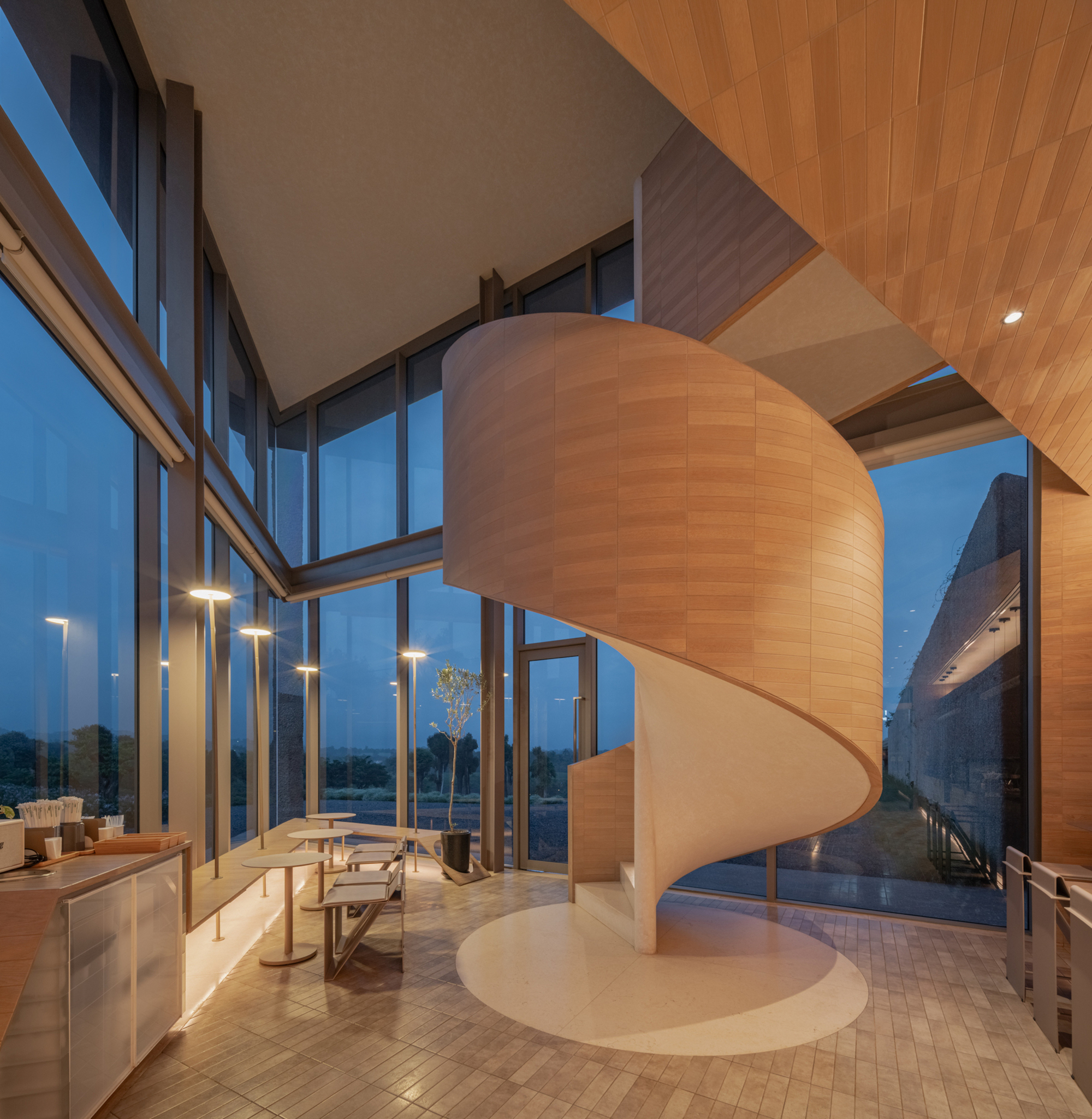 Timber-clad helical staircase rising from herringbone floor beside full-height glazing at twilight