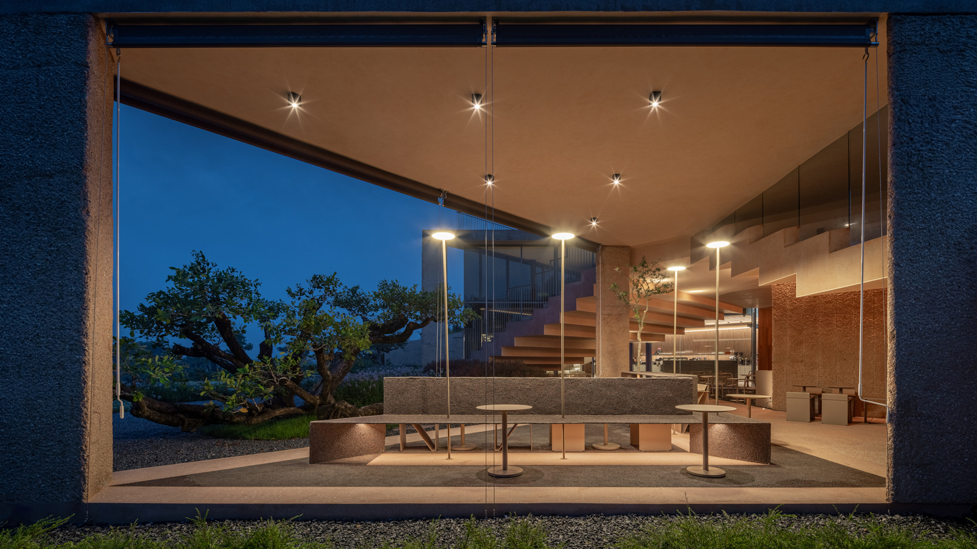 Glass-enclosed seating pavilion with pendant lights and view to trees at night