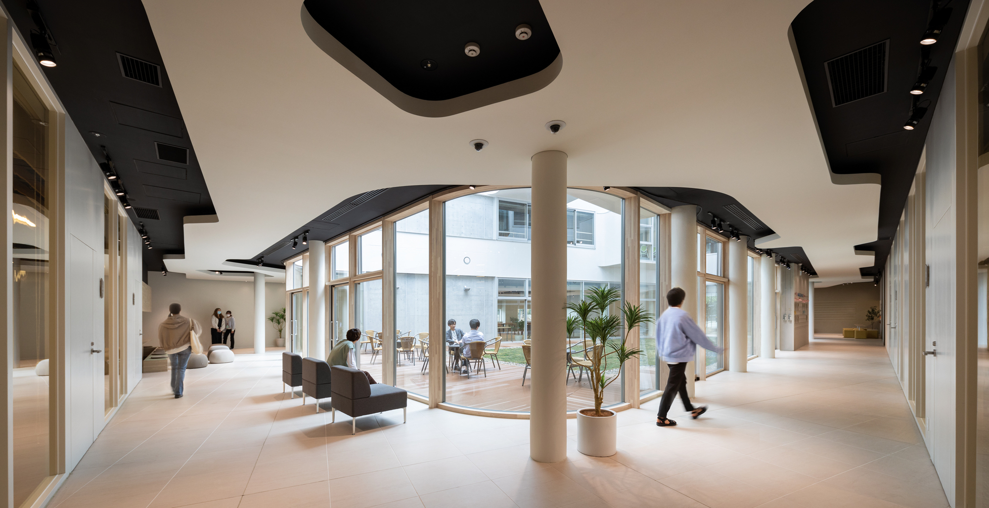 Interior corridor with curved glazed wall, black ceiling panels and cylindrical column as people walk through