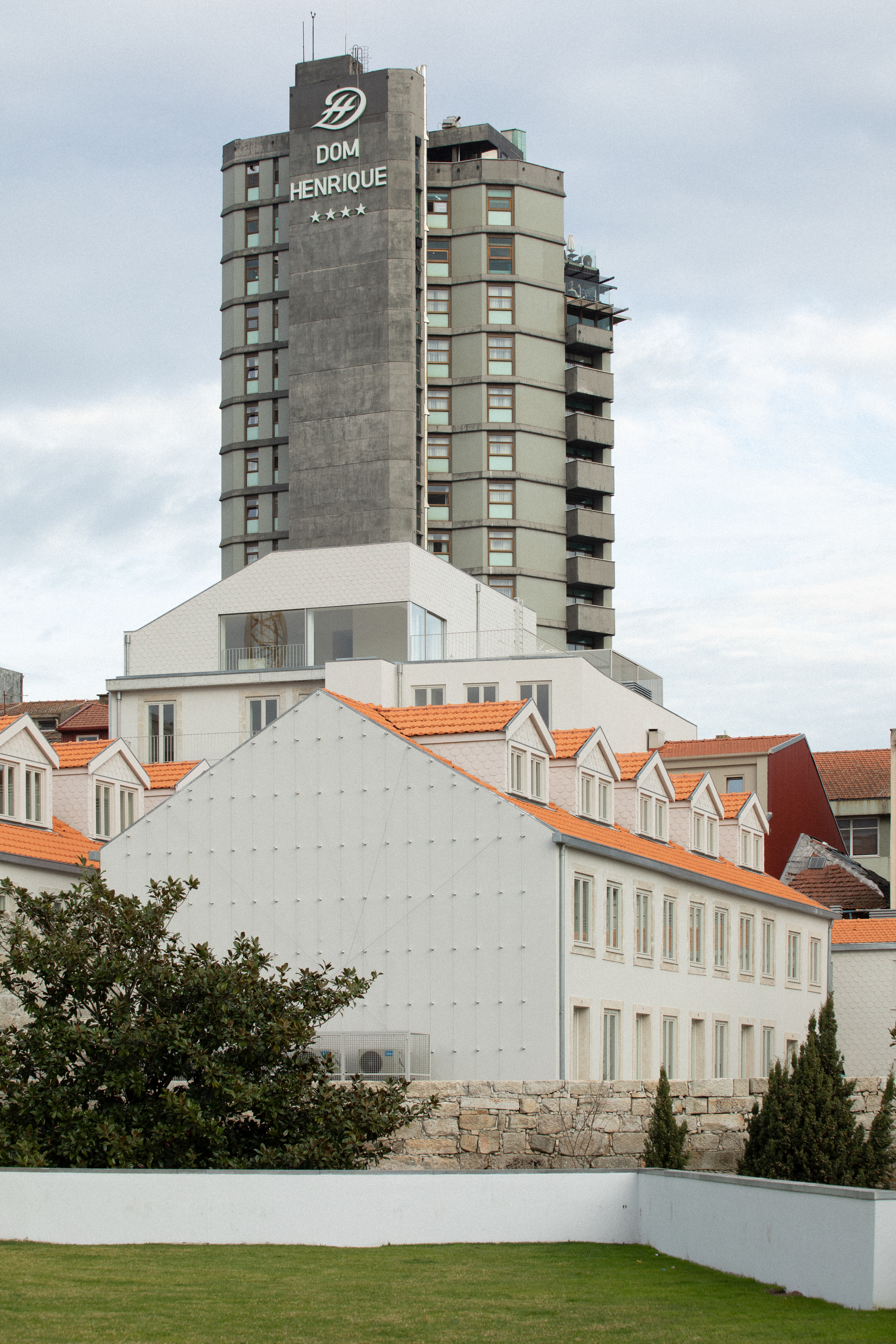 White gable facade and orange-tiled roofline beneath a concrete high-rise tower under cloudy skies