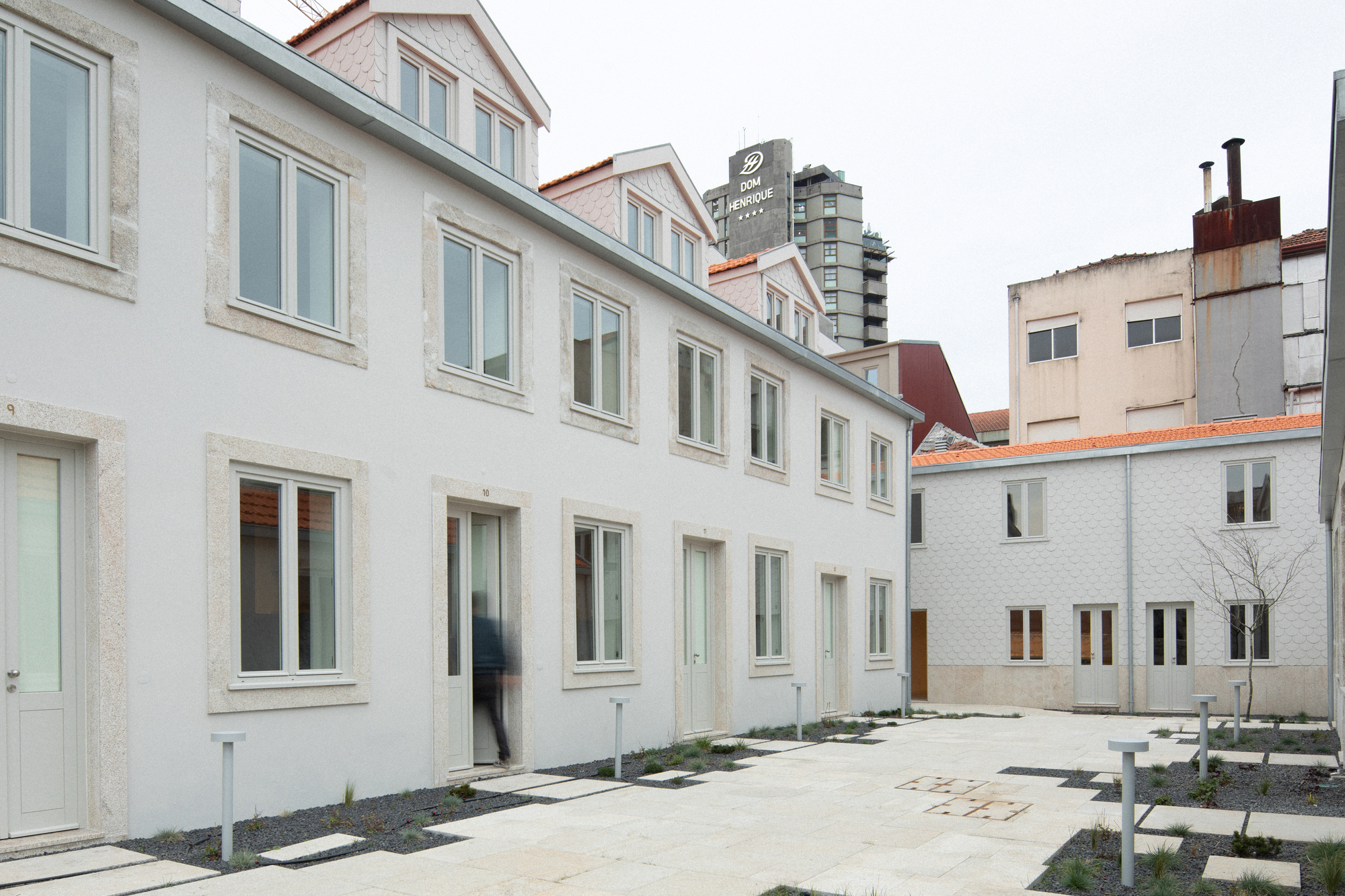 Paved courtyard between white-rendered residential facades with scattered planting beds and young saplings