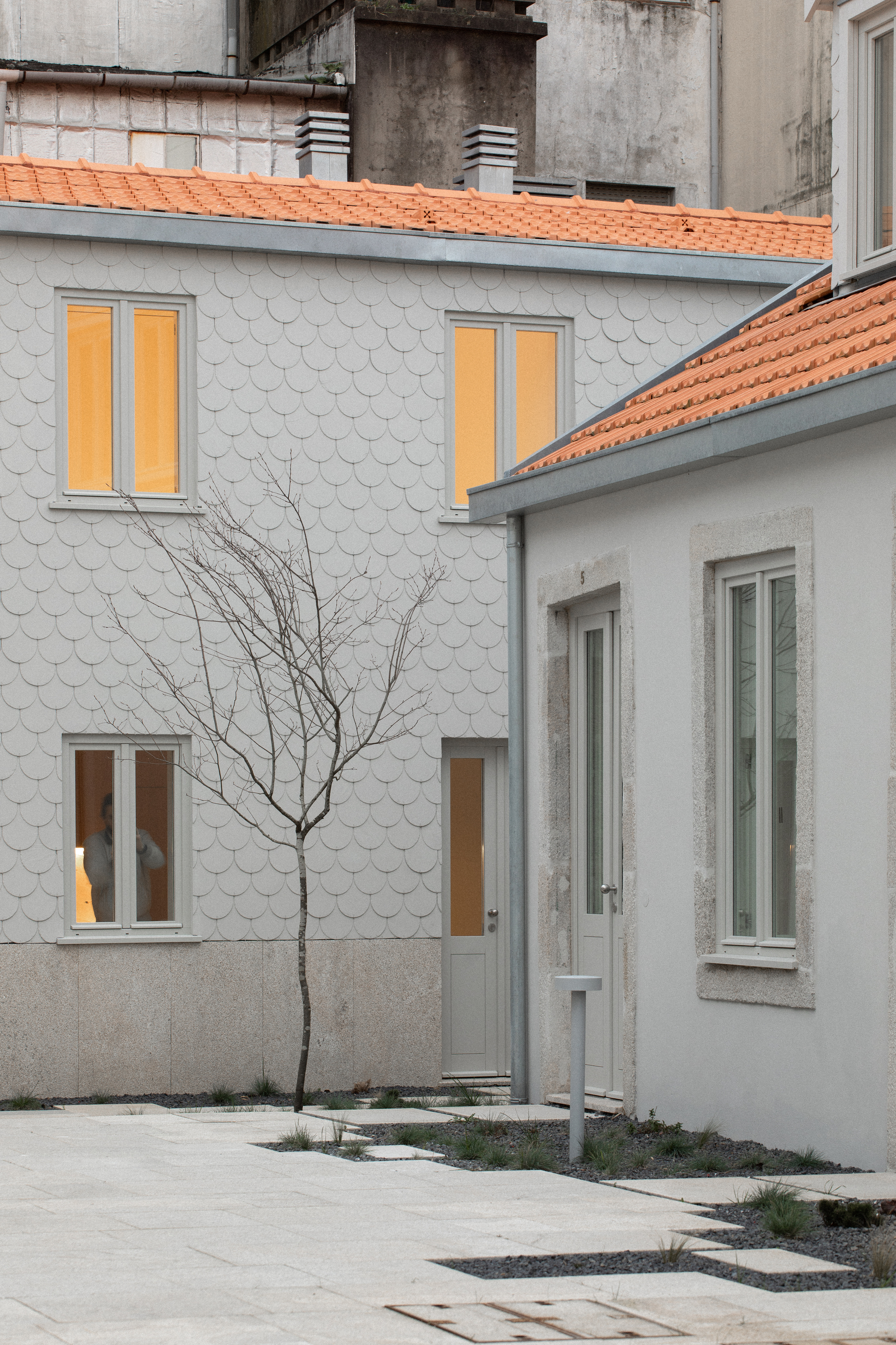 Facade clad in white scalloped tiles with double-hung windows and terra-cotta roof beside a bare winter tree