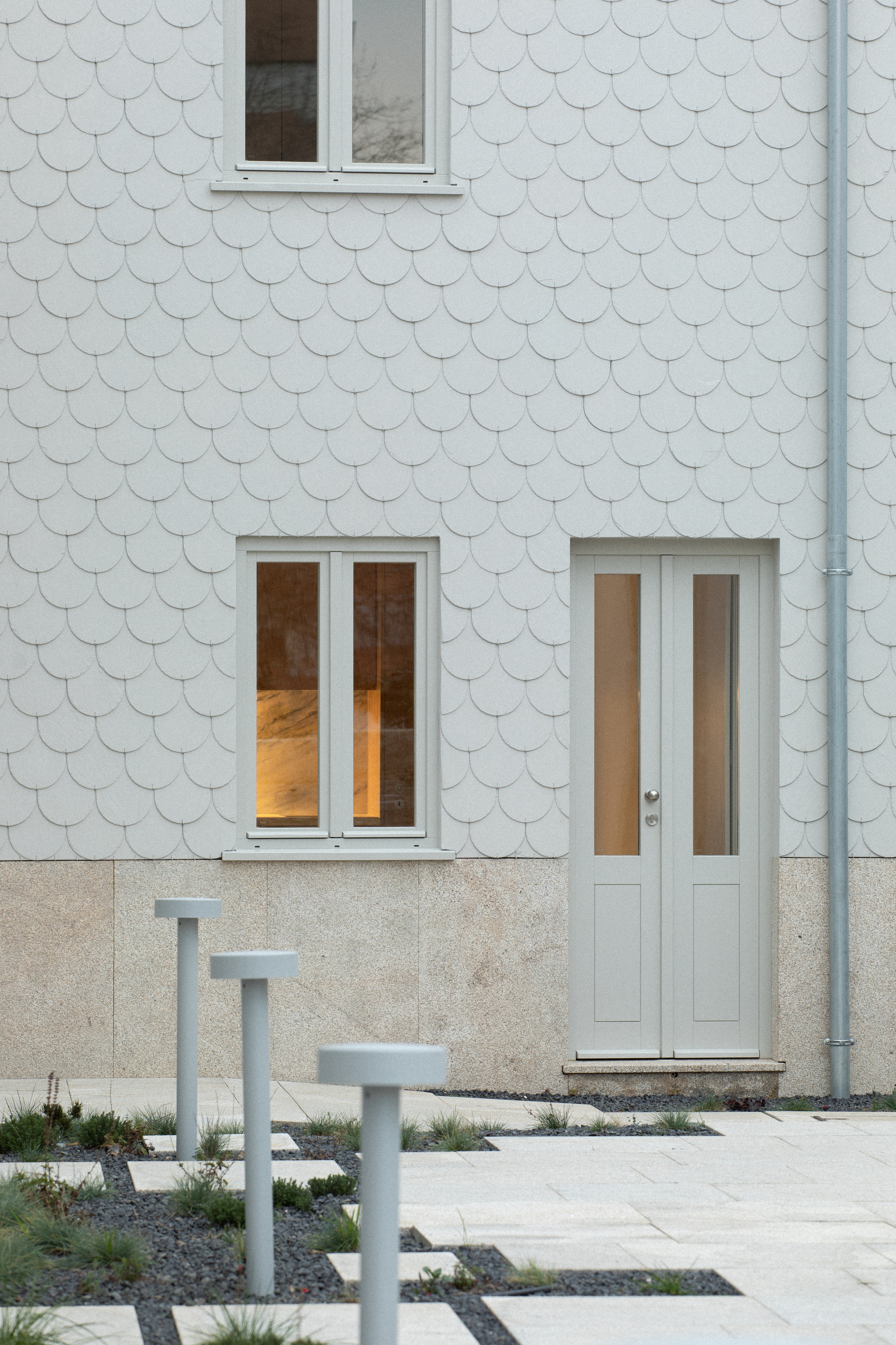 Close-up of scalloped tile cladding above a granular base with steel posts punctuating the paved ground plane