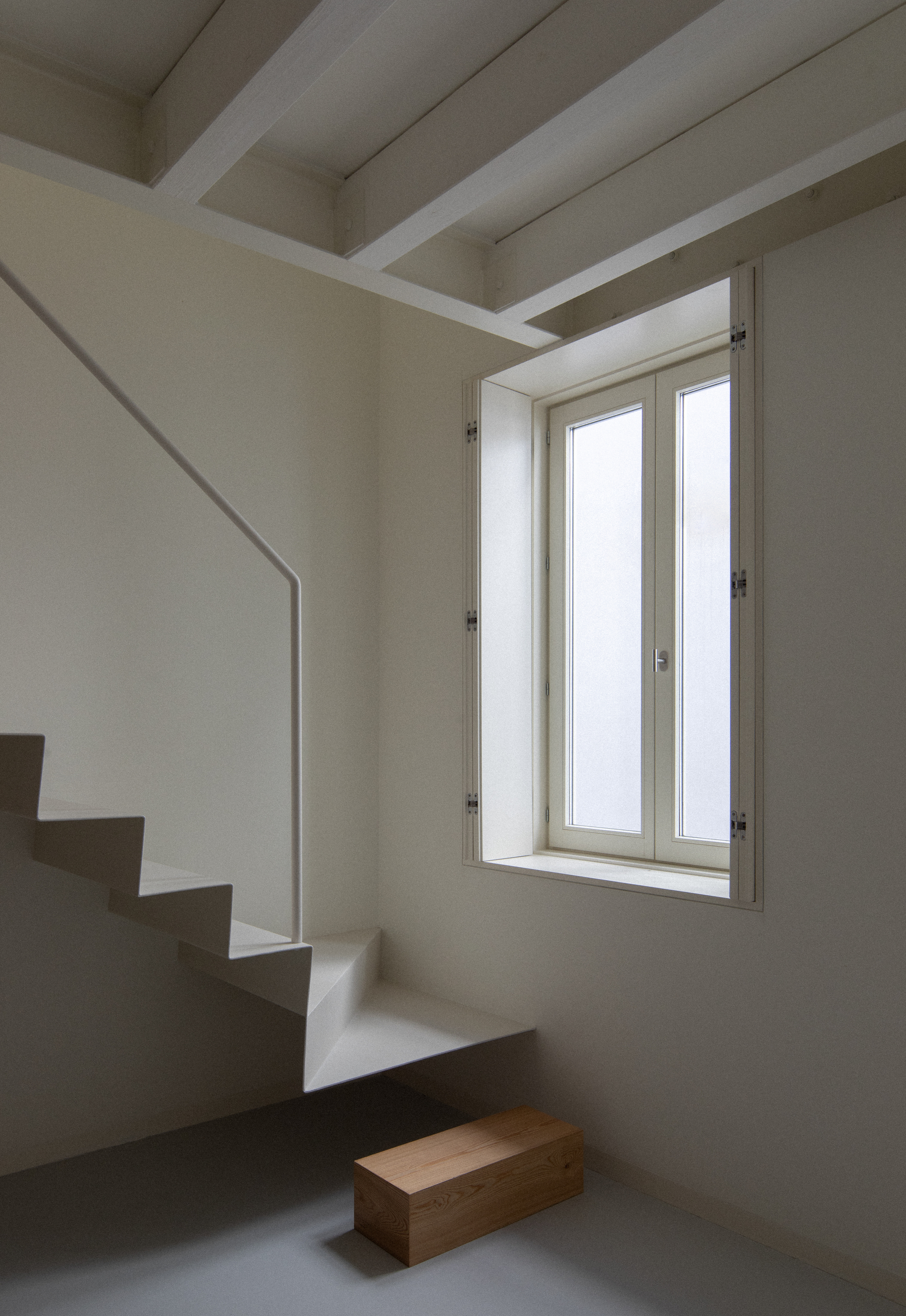 Interior staircase in white plaster with exposed ceiling beams and a casement window casting afternoon light