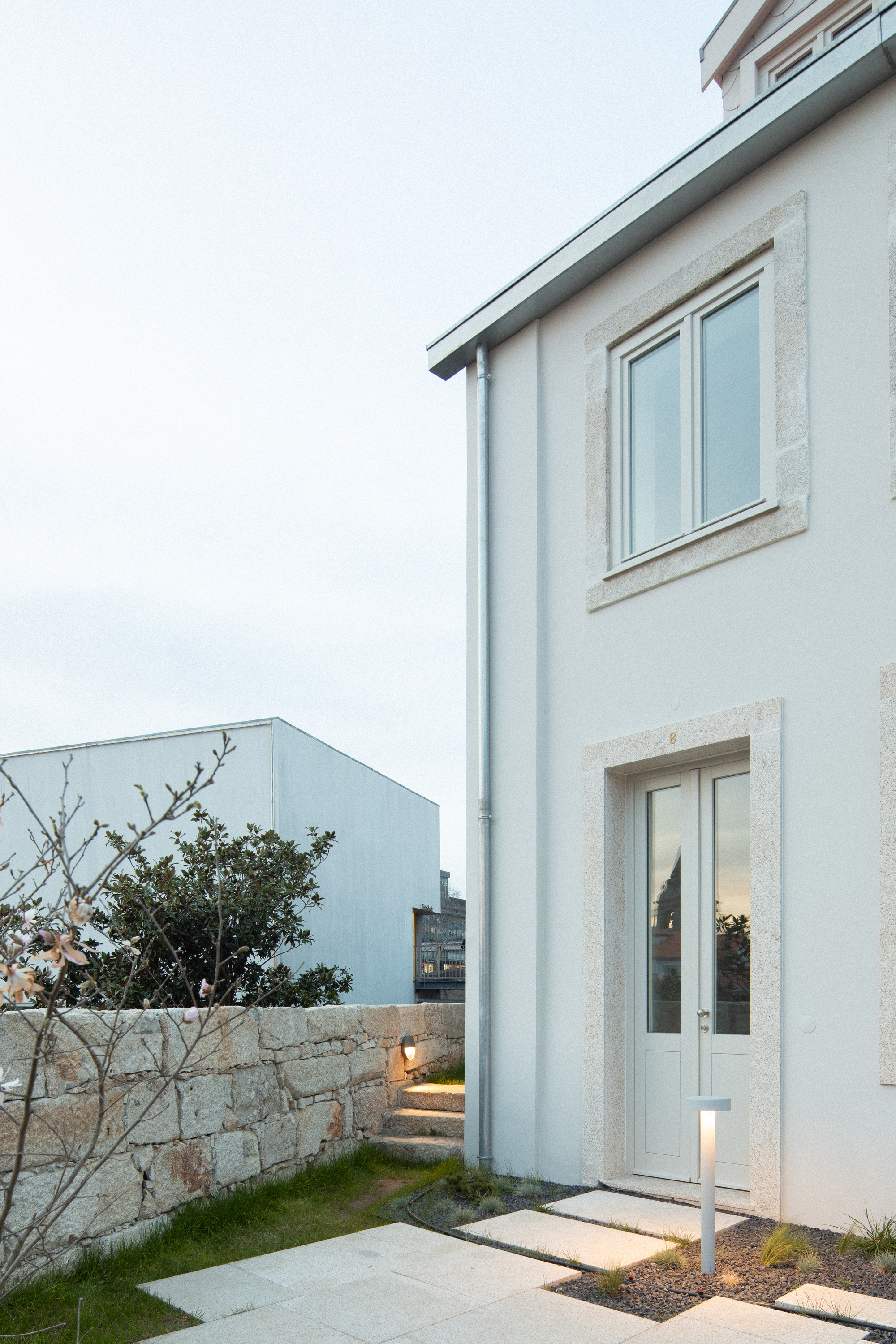 White rendered facade with stone window surrounds beside a low granite wall at dusk