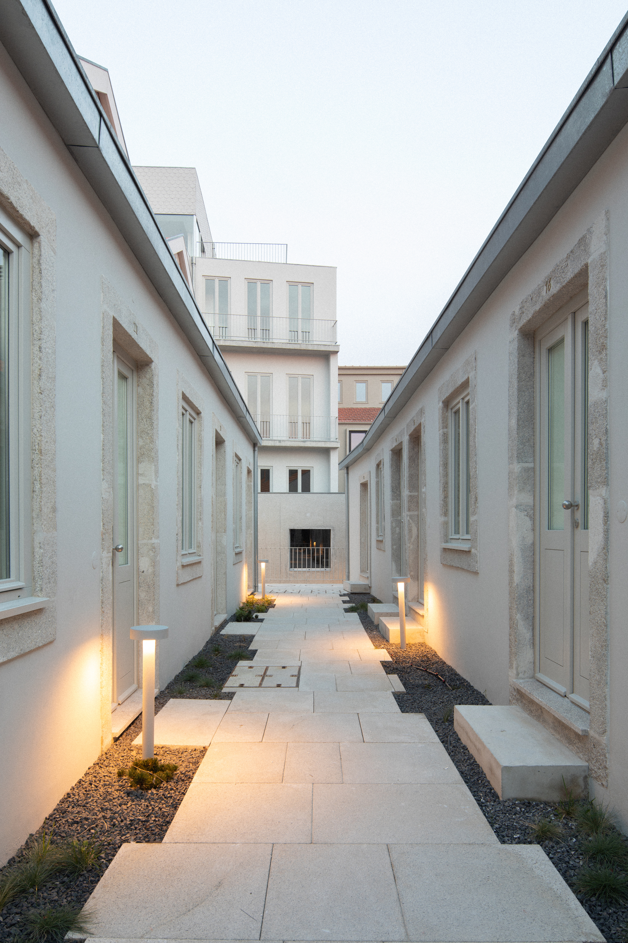 Linear courtyard with pale stone paving and uplighting between two white rendered blocks at dusk