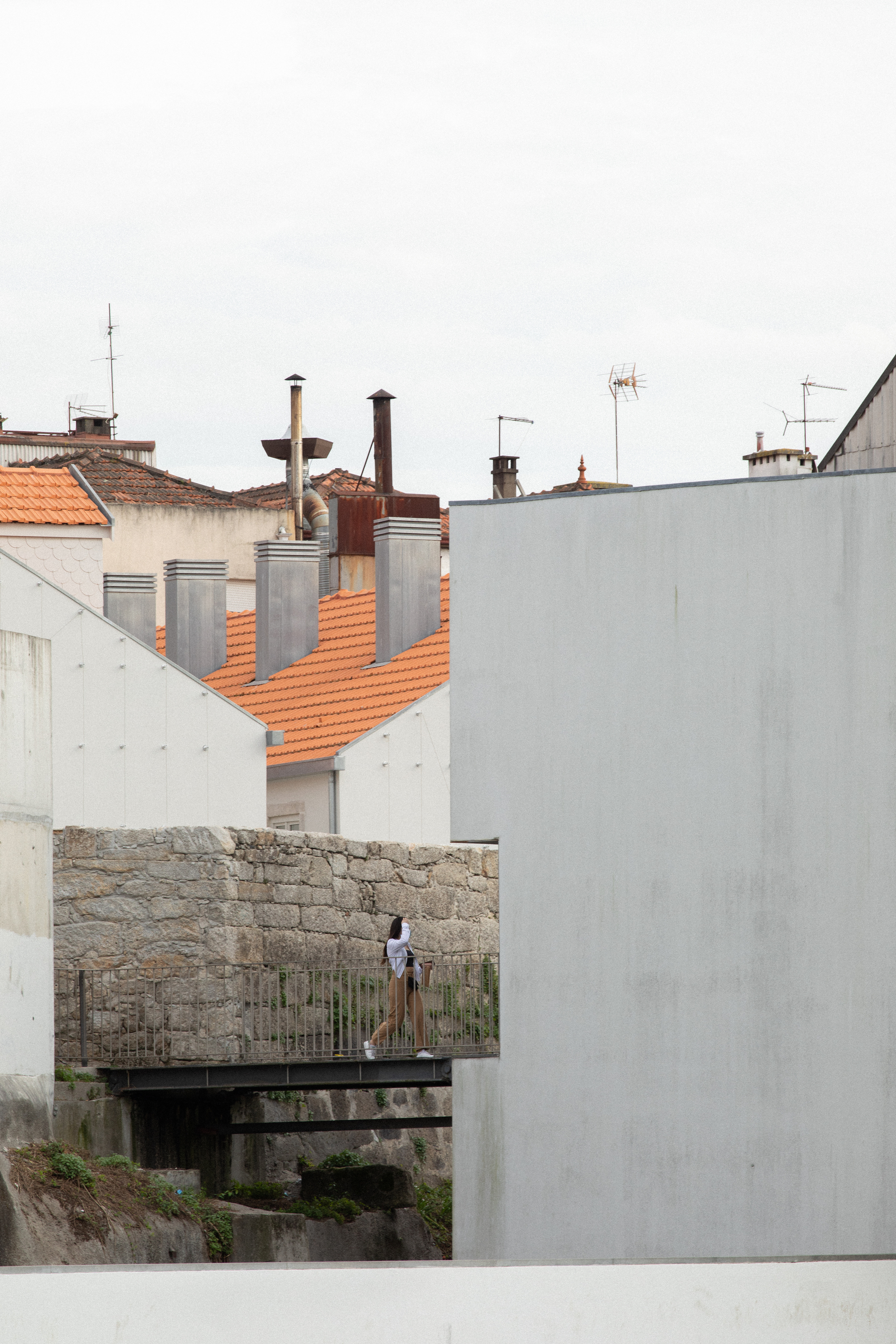 Figure standing on a timber footbridge between white concrete walls and terracotta rooftops