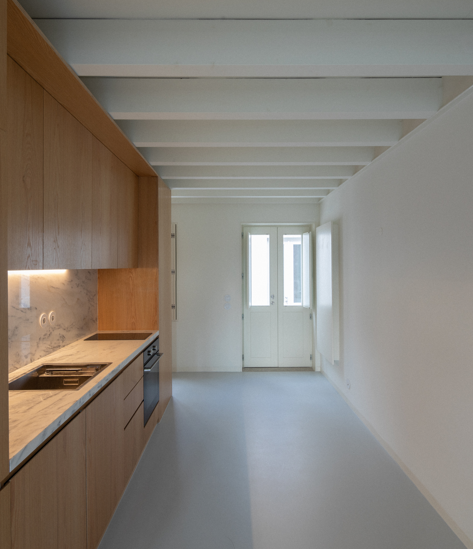 Narrow kitchen with oak cabinetry and exposed ceiling beams leading to a white double door