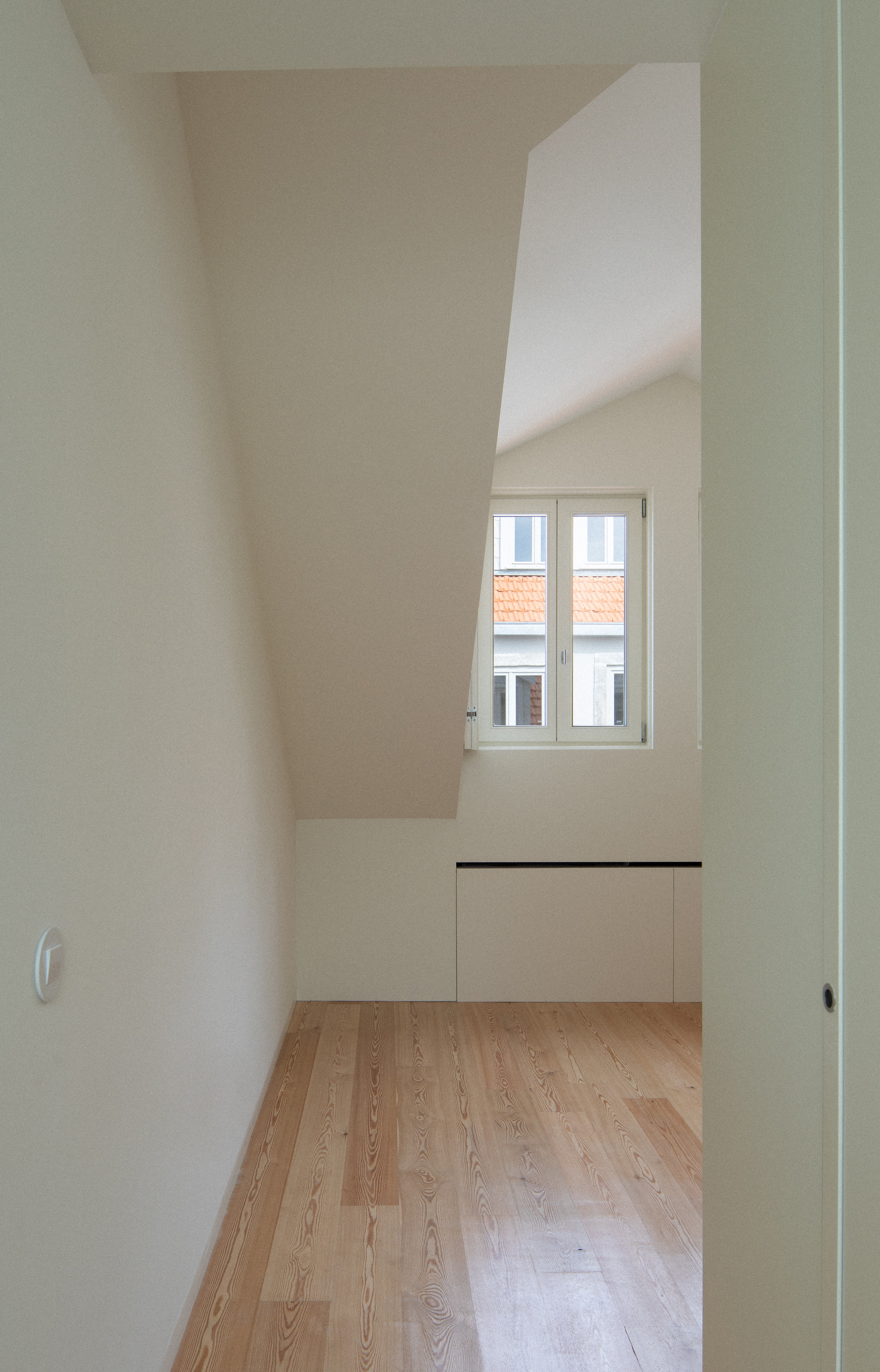 Small bedroom with oak flooring and low built-in cabinet below a window with rooftop view