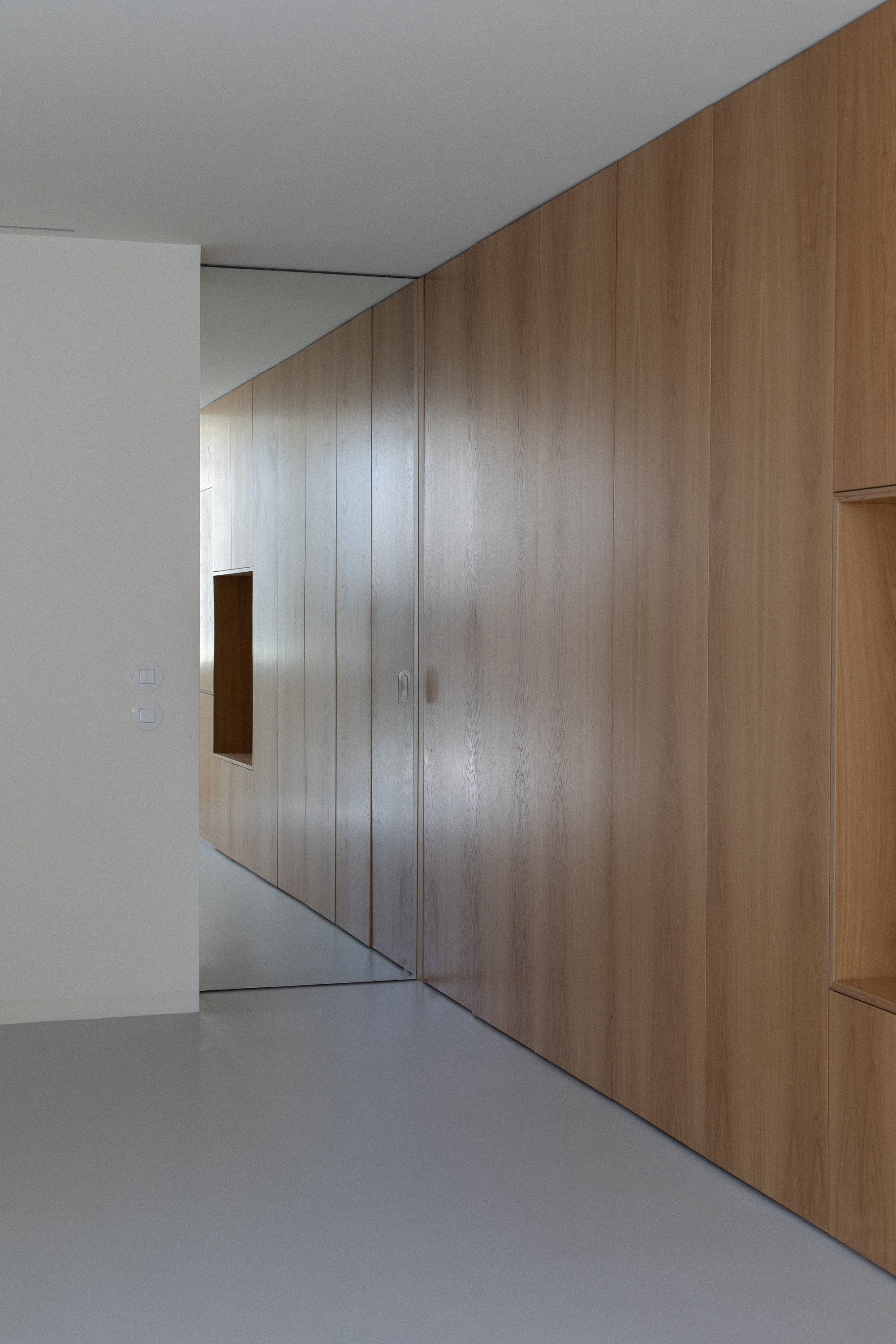 Angled oak veneer wall with small square opening above grey resin floor
