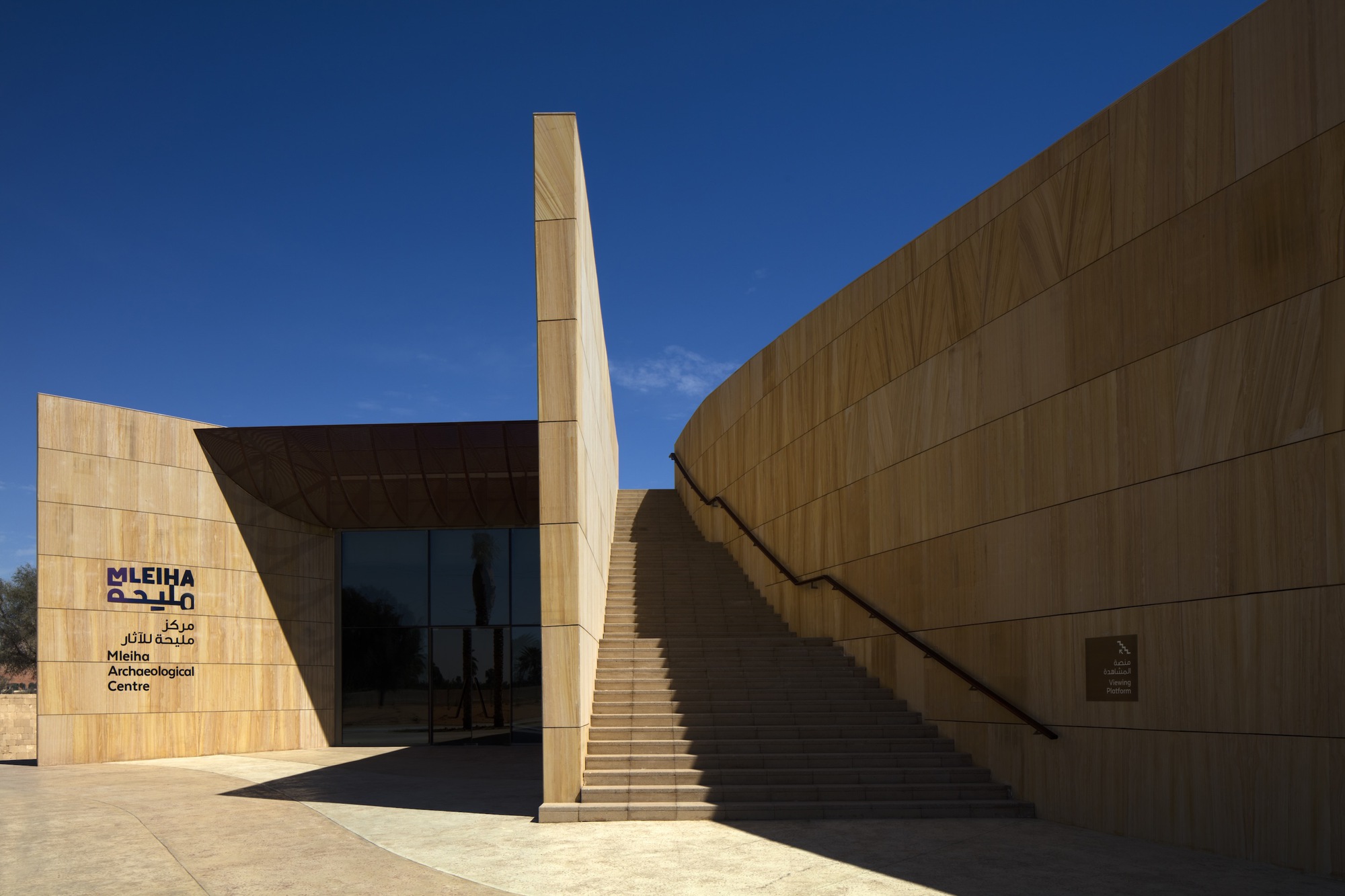 Entrance plaza showing sandstone-clad facade, freestanding tower element, and exterior staircase to roof