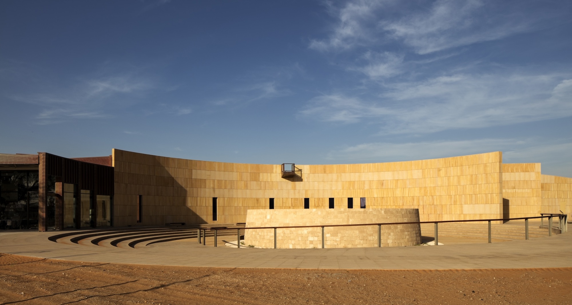Curved sandstone facade with narrow vertical windows and stepped amphitheater seating under a blue sky