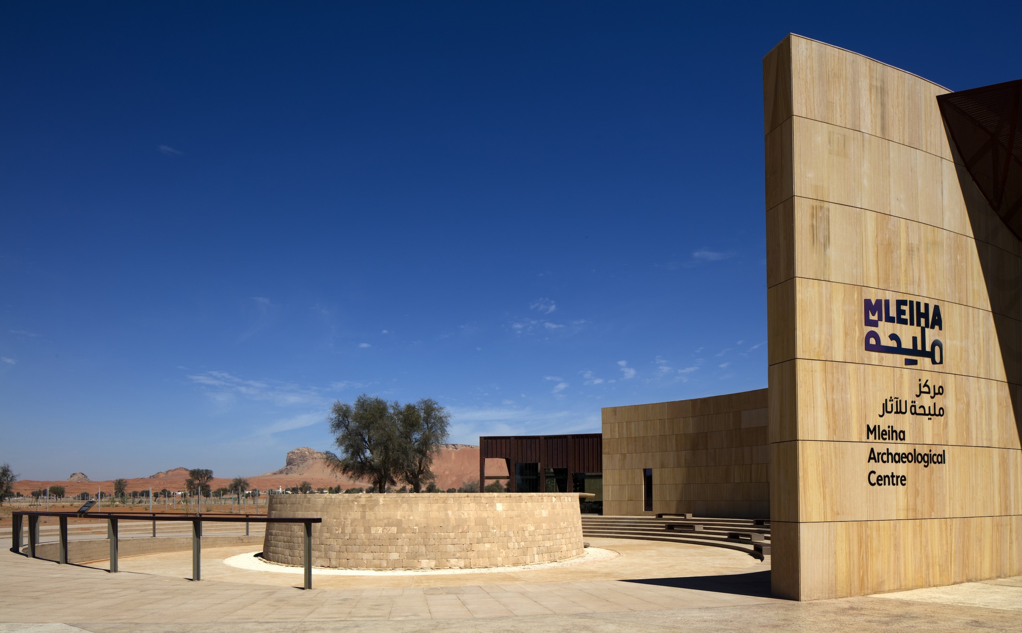 Wood-clad entrance tower with signage beside a curved stone wall and desert landscape beyond