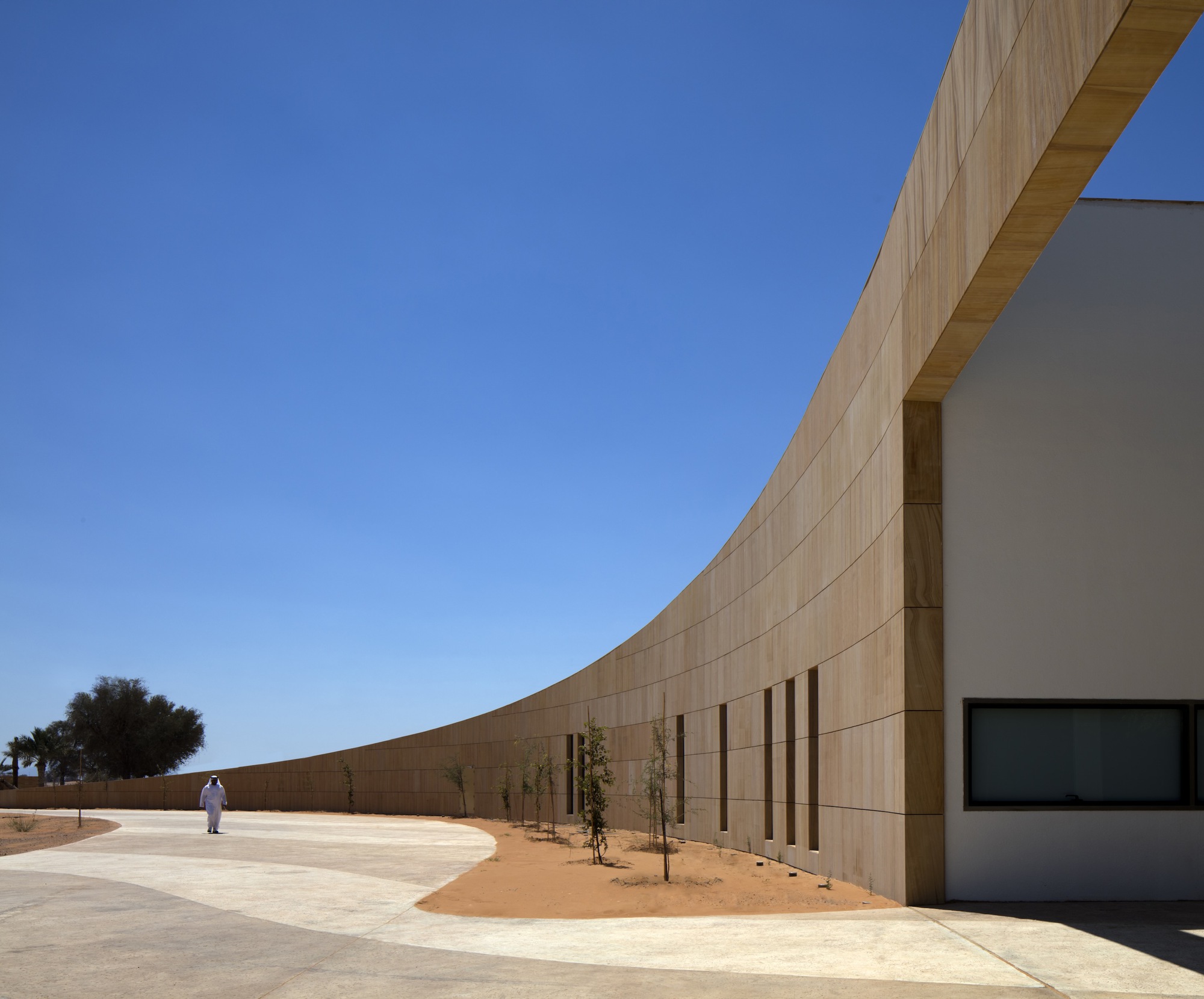 Curved sandstone wall with vertical window openings and a figure walking along the pathway