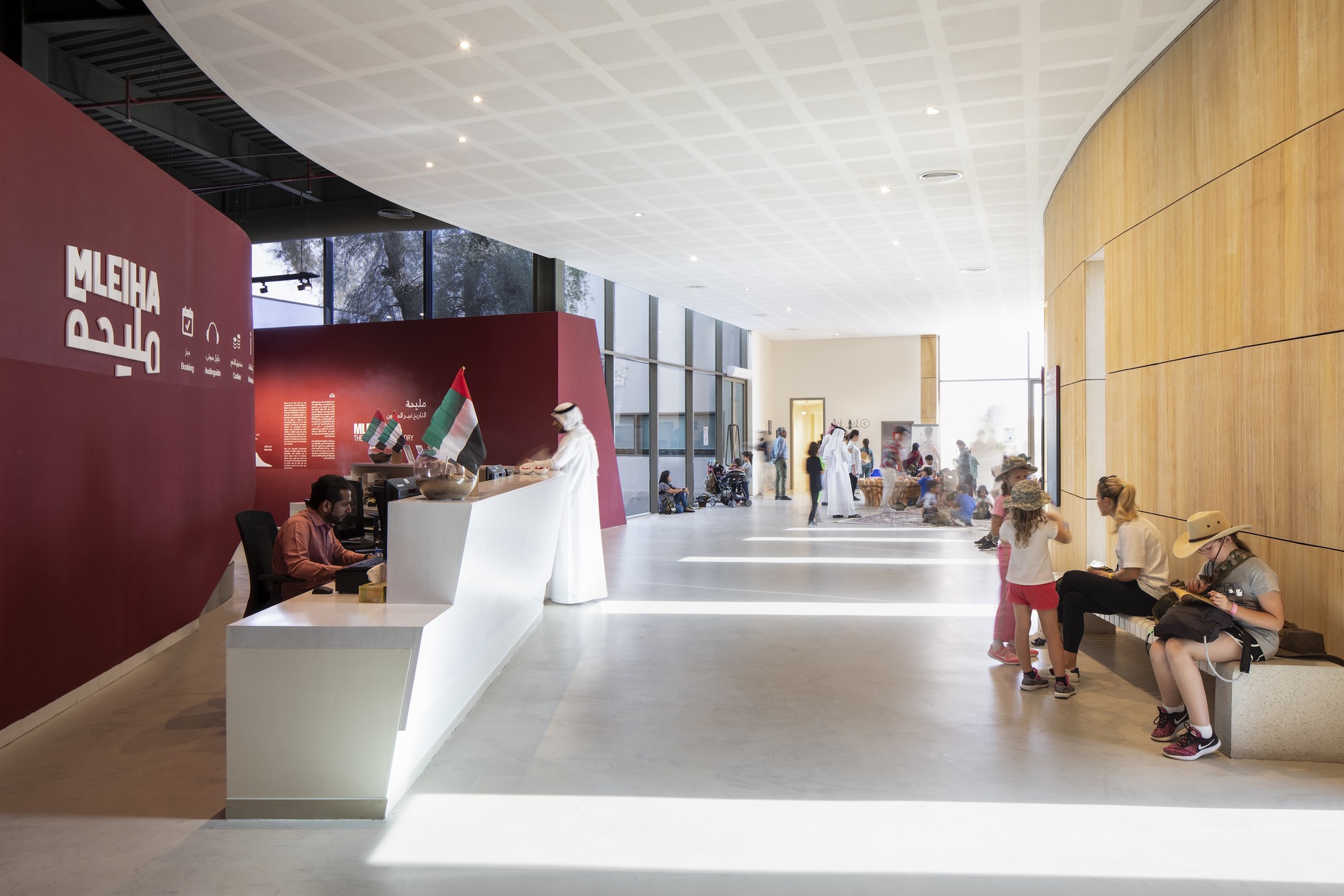 Reception hall with white desk, curved wood wall, and visitors seated on benches in daylight